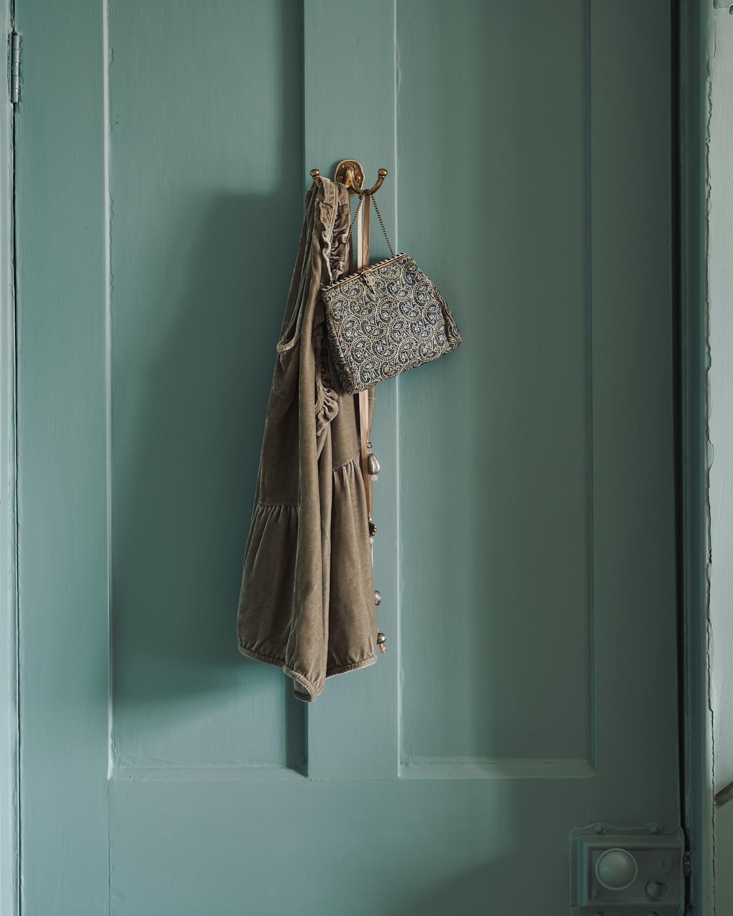 A Parisian bohemian-style twin brass hook, fitted to a blue painted door. Glamorously holding onto a lovely blue detailed vintage bag and an olive green suede light jacket.