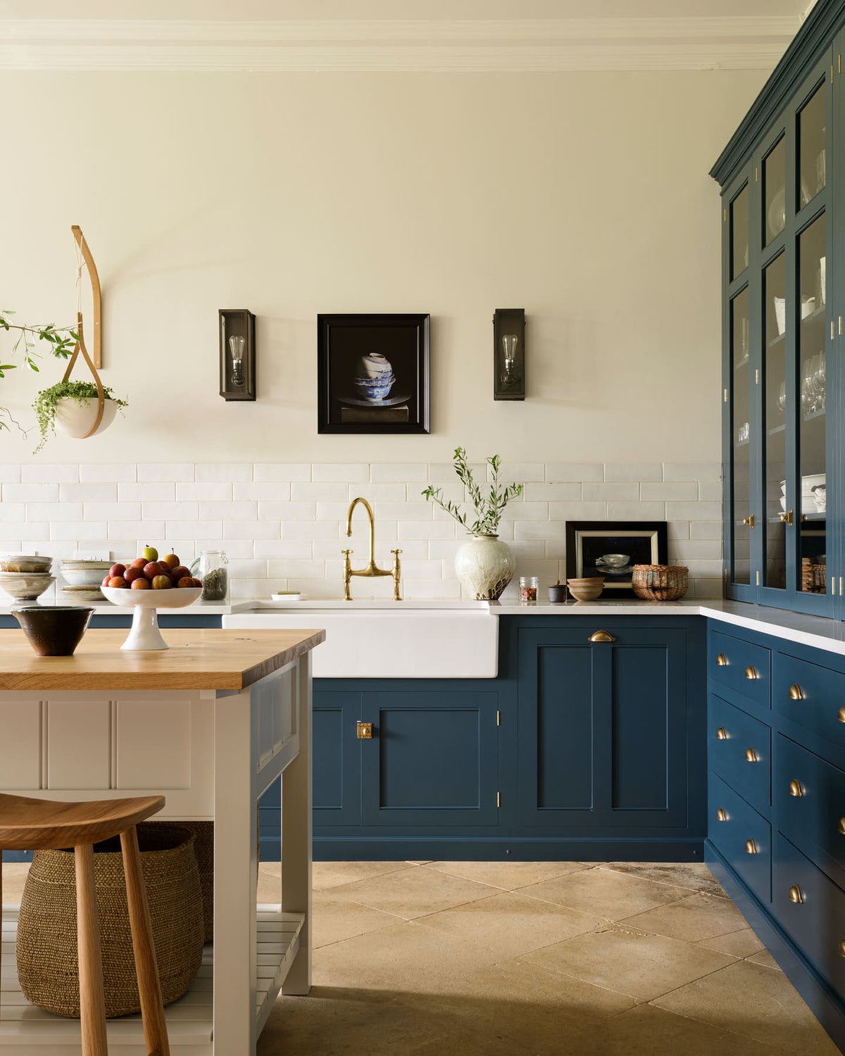 A classic English, Bond Street blue kitchen features a white linen prep table with a rustic oak worktop, a cooker and sink run with a silestone worktop, glazed counter cabinet, double sink, an aged brass Ionian tap, and white crackle metro splash back tiles. The renovated medieval house is elegantly adorned with modern still-life paintings, artisan ceramics, and vibrant green plants, seamlessly blending the historical with the contemporary.