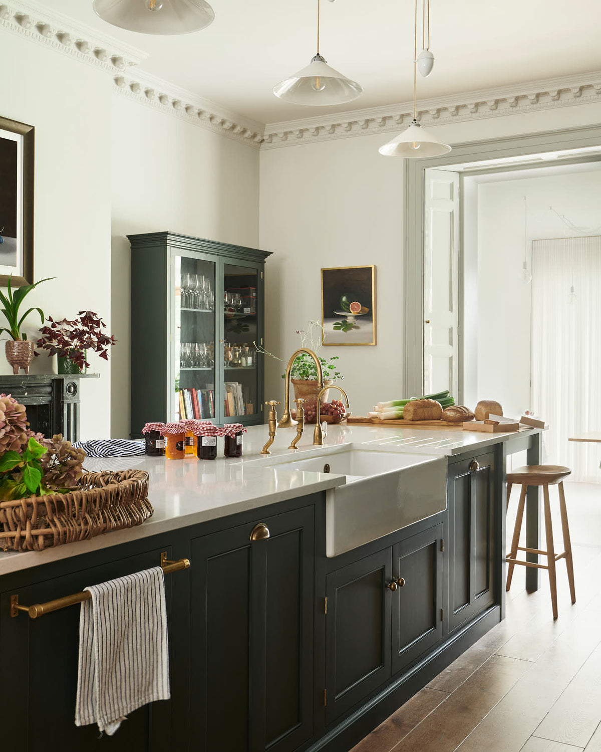 An oblique view of a long kitchen island with a double sink, an aged brass Ionian tap, and a curiosity cupboard in the grand bakehouse green classic English kitchen. Adorned with loaves of bread, leeks, red grapes, potted plants, still life paintings, and homemade jams; the room feels homey and welcoming despite its large and decorative appearance.