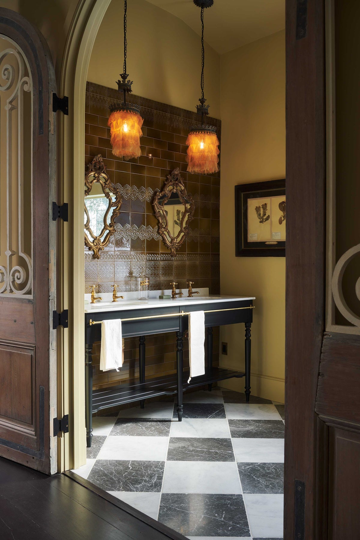 Looking past the antique Parisian doors into a beautiful butter-yellow powder room, you’ll find a black twin Victorian washstand, tall sepia lace market tiles, and a Parisian chequered marble floor. Paired with antique mirrors, pendant lights, and framed pressed botanical artworks, the room exudes a glamorous, vintage charm.
