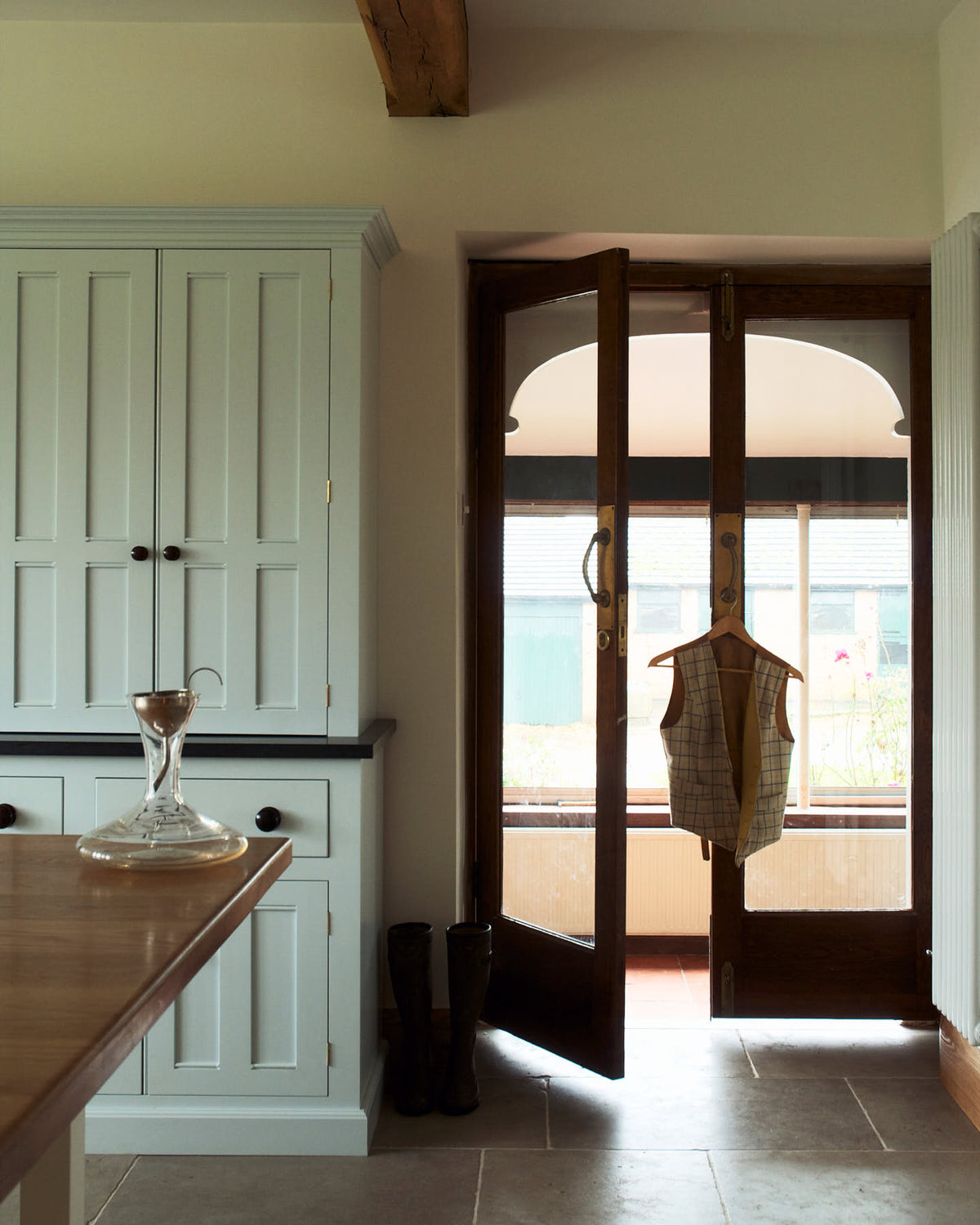 This simple and spacious classic English kitchen features a kitchen island with a solid oak worktop, a double Belfast sink, an antique silver Ionian tap, and a free-standing closed larder pantry cupboard. Adorned with a glass decanter featuring a metal funnel and a vest hanging on the door handle, this kitchen creates a humble and stylish space.