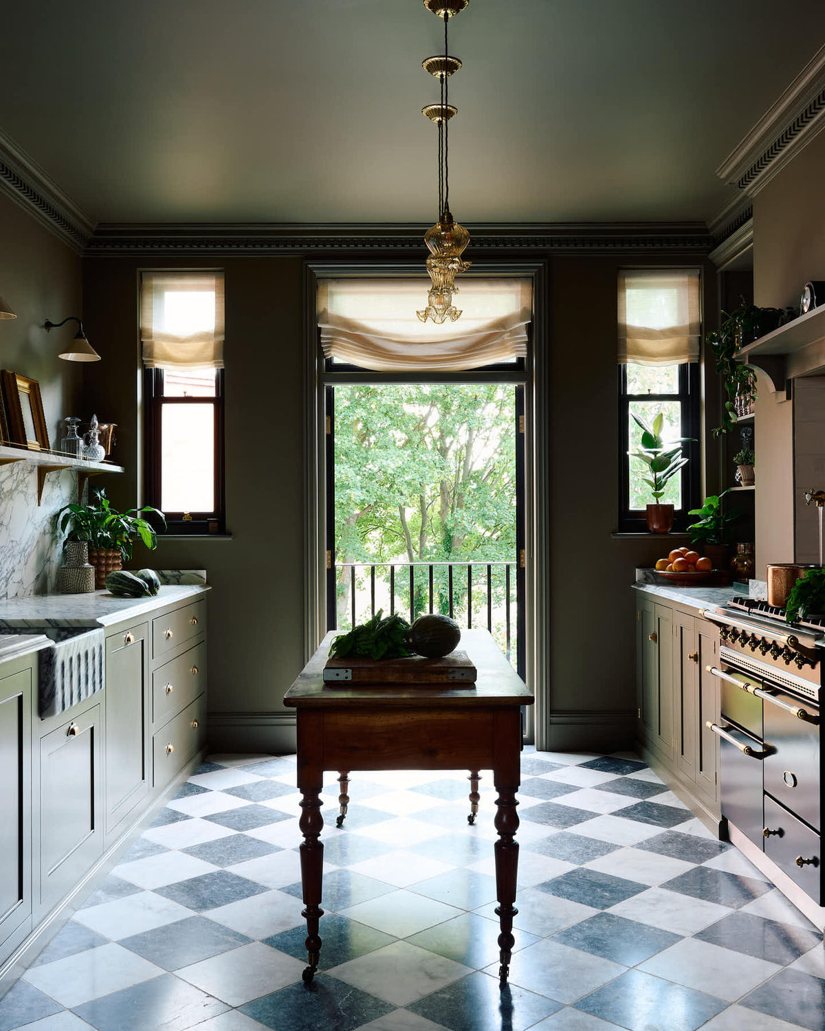 A grand and atmospheric olive green classic English kitchen features a marble worktop and splash back along the sink run, with a single marble sink, an aged brass Ionian tap, and an antique prep table on castors. Grand chequerboard marble tiles lead out onto a balcony overlooking green trees.