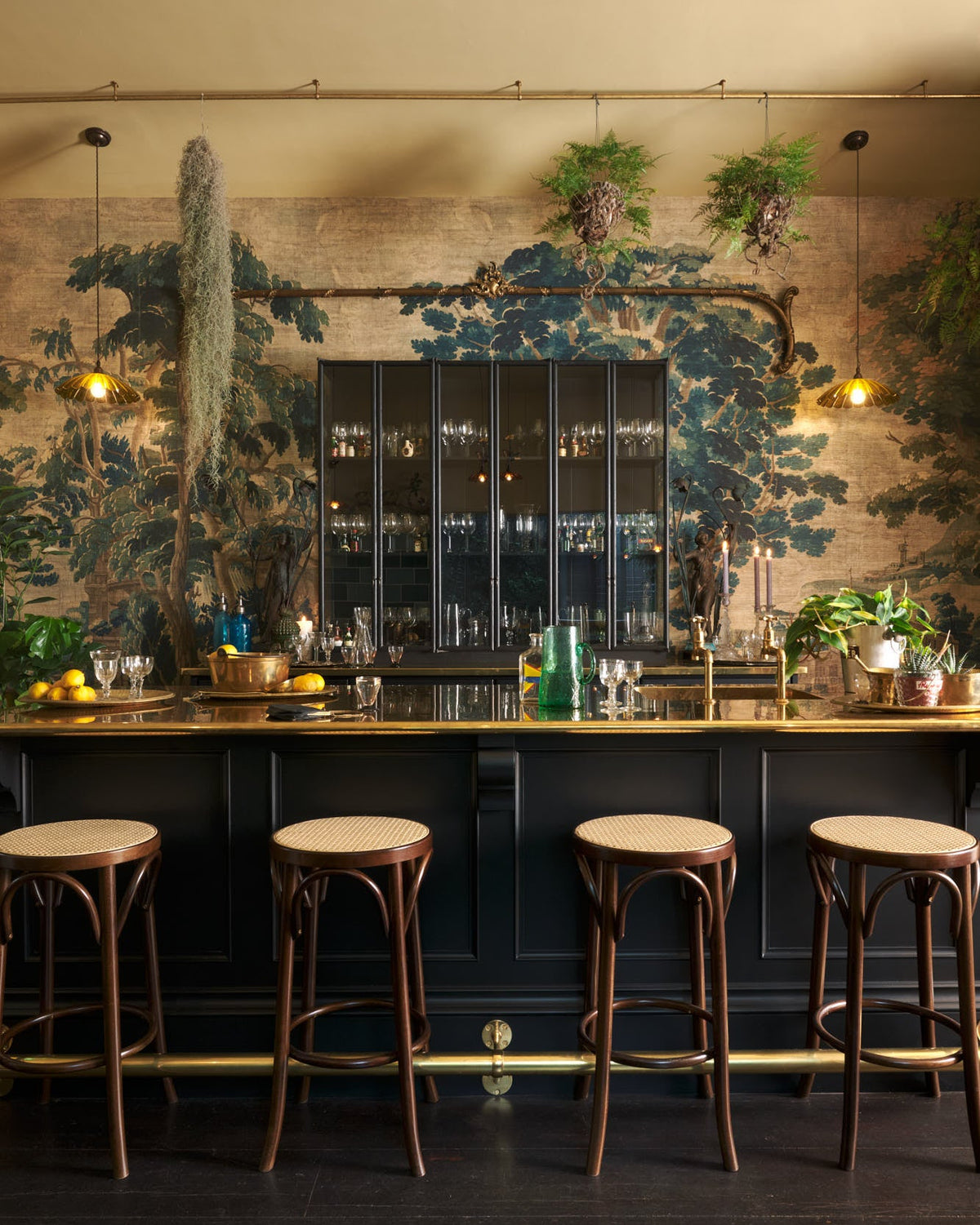 A chic and glamorous Bobbin Black Haberdasher’s bar featuring a kitchen island with a polished brass worktop, a single polished brass sink, aged brass Mayan taps, a brass foot rail, glazed base cabinet with internal drawers, tall countertop cupboards, and woven top bentwood café stools. Set against an illustrated foliage wallpaper and adorned with hanging plants, wine glasses, and vintage decanters, it creates a beautiful and intimate personal bar.