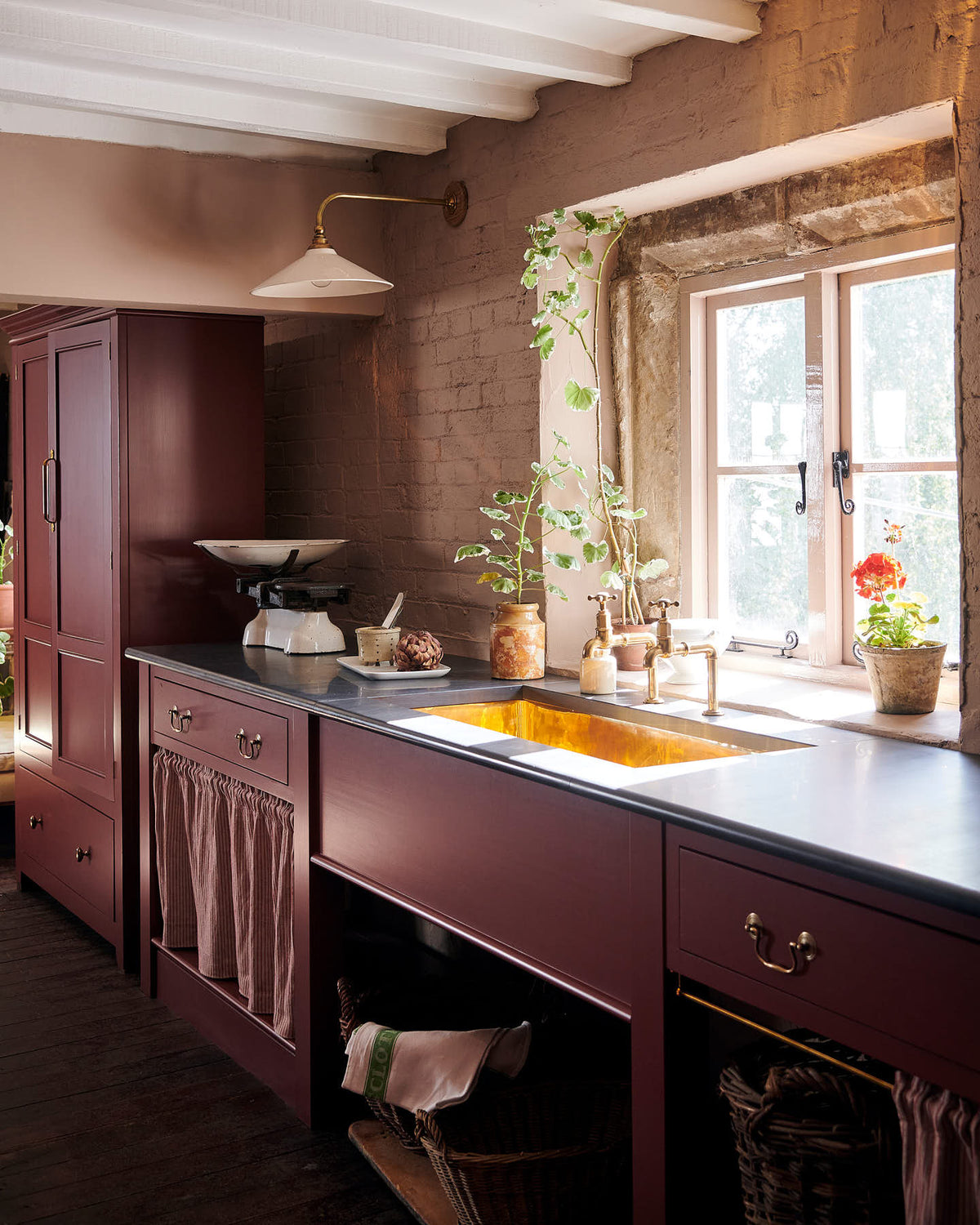 A closer view of a refectory-red Heirloom kitchen’s sink cabinet reveals a polished brass sink, aged brass Mayan taps, café curtain rails, a red and white striped curtain, and a pantry cupboard. Beautifully decorated with an Heirloom task light, a vintage scale, artisan ceramics, and house plants, this kitchen offers a modern twist on vintage furniture, creating a classic space designed to last.