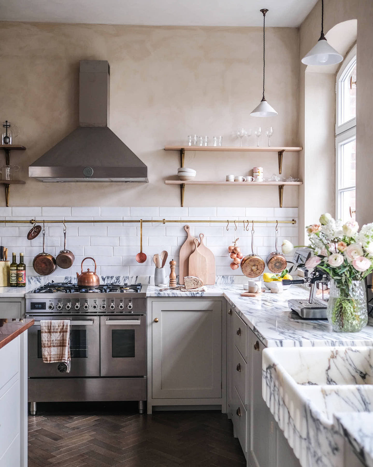 A grand yet grounded shaker kitchen in muted mushroom off-white, alive with scattered pots, pans, fruits, and vegetables. The L-shaped cooker and sink run features an Arabescato marble countertop, double-fluted sink, and aged brass Ionian tap. Crackle metro tiles and a long-aged brass rail form the splash back. A large copper-topped island and cosy breakfast nook complete the space.