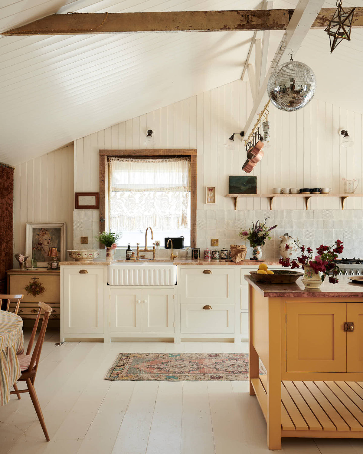 A bohemian kitchen with cream coloured Shaker-style cupboards, floorboards and walls. The room is decorated with an assortment of vintage pieces of furniture, paintings and fabrics.