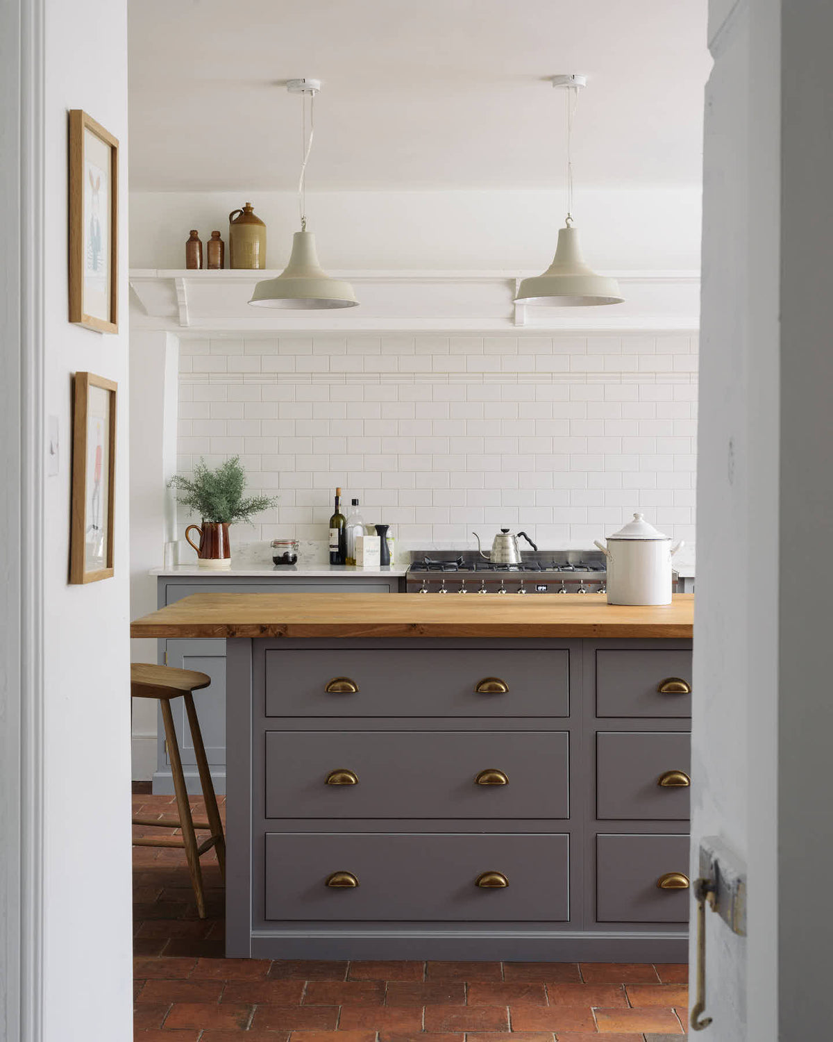 Looking through a narrow hallway, a simple, modern shaker kitchen in a historic Cheshire townhouse, featuring a silestone worktop cooker run, and a large island with a solid oak worktop and a breakfast nook. Painted in muted cool grey with white walls and warm terracotta tile flooring.