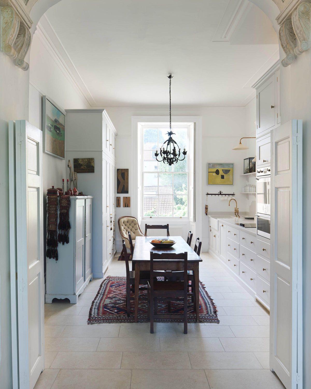 Through a grand decorative archway is a tall yet simple Shaker kitchen featuring a sink and cooker run with a Carrara marble worktop, shelves, and a small bank of larder. Lovingly decorated with antique furniture, a vibrant red decorative rug, a variety of chic and curated paintings, and a vintage metal chandelier.