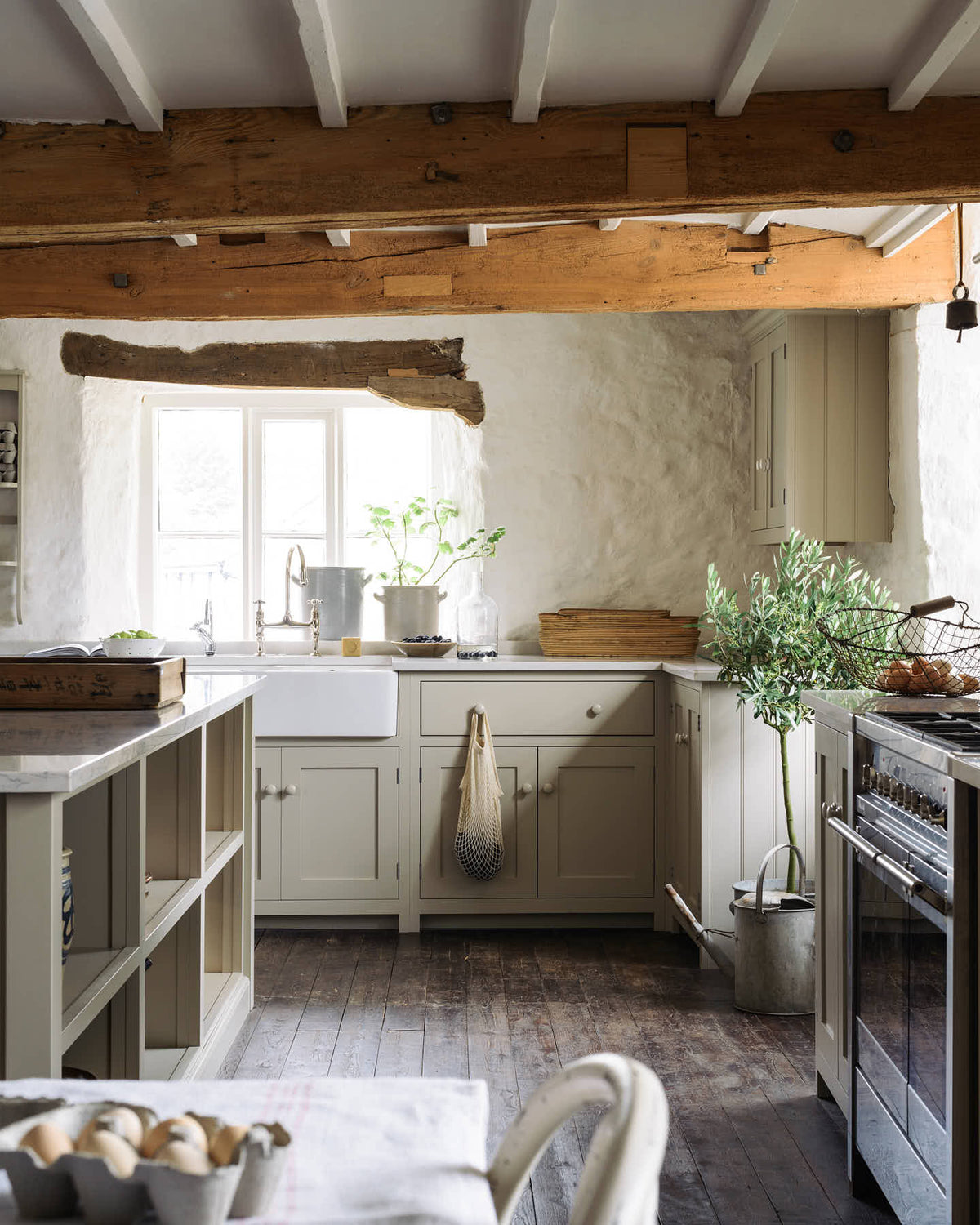 A sink run in a simple English farmhouse kitchen at deVOL’s headquarters, Cotes Mill. Low-exposed wooden beams and worn floorboards create a rustic atmosphere, while textured off-white walls add charm. The Shaker kitchen, including its island, is painted in Mushroom—a soft, timeless beige.