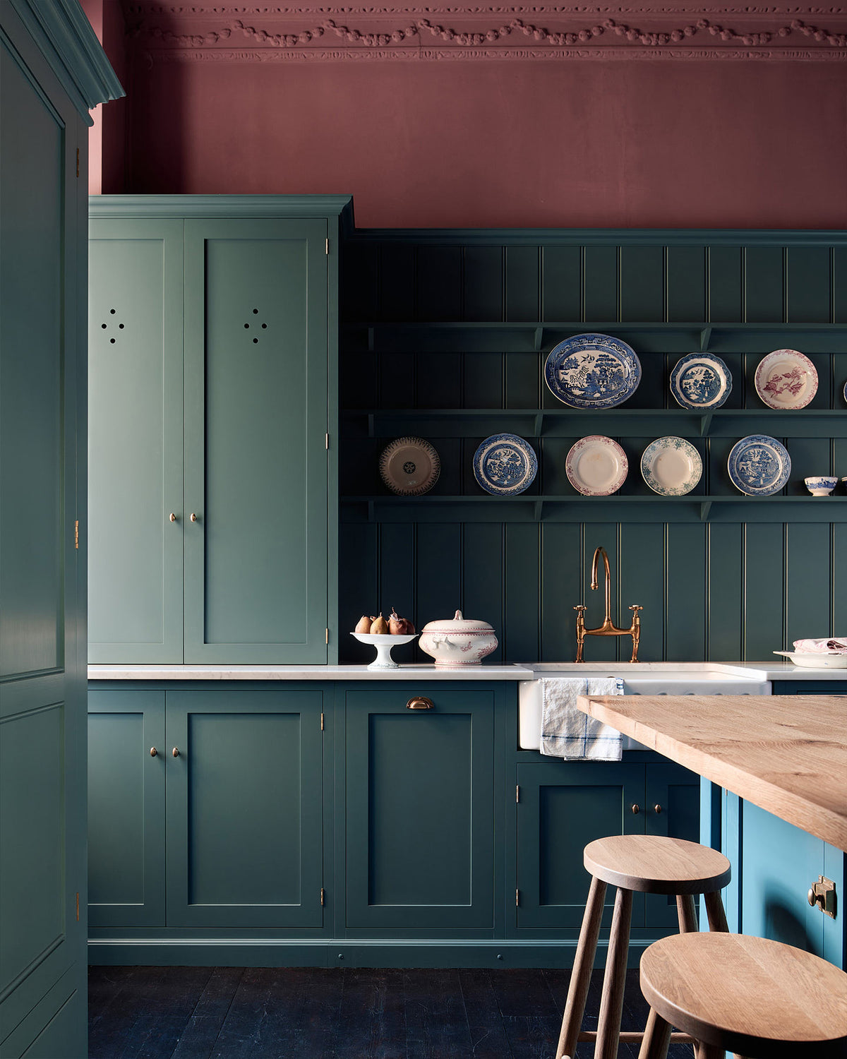 A shaker kitchen’s sink run with a countertop cupboard and panelled wall beside an original sash window. A small island with a rustic oak worktop stands at the centre, accompanied by a row of understated oak Helen’s Stools. Behind them, a grand freestanding pantry from our Classic English range is accented with Victorian-style brass Butler’s Knobs.