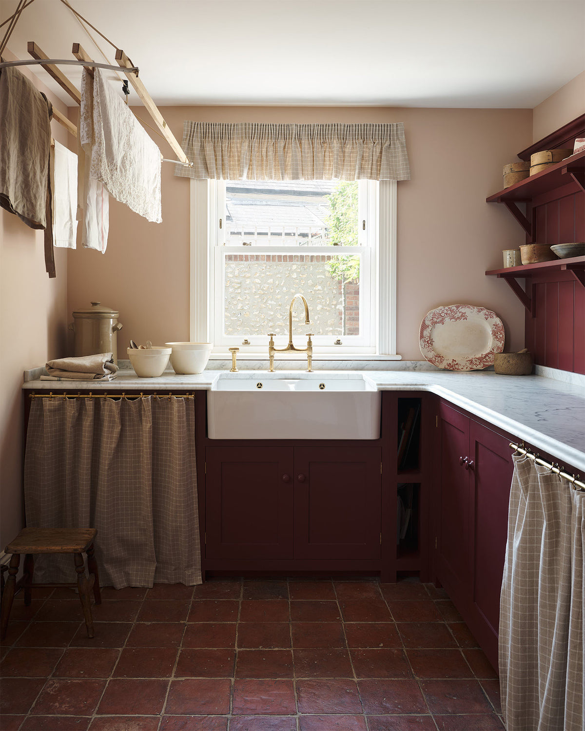 An L-shaped laundry room with fitted Shaker base cabinets painted in a dark, berry-red shade and linen curtains hanging from aged brass rails to hide the appliances. A wooden laundry maid and rustic terracotta tiled floor give the space a utilitarian feel, whilst delicate pink walls and honed marble worktops add a little bit of luxury.