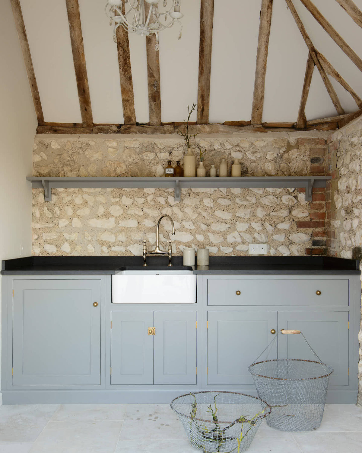 A chic and simple muted-blue sink run against the original wall textures of the pool house features a single Belfast sink with a nickel Ionian tap, a black honed granite worktop, and a long shelf. Adorned with vintage ceramics, brass fittings, and vintage steel baskets, it creates a functional corner in the space.