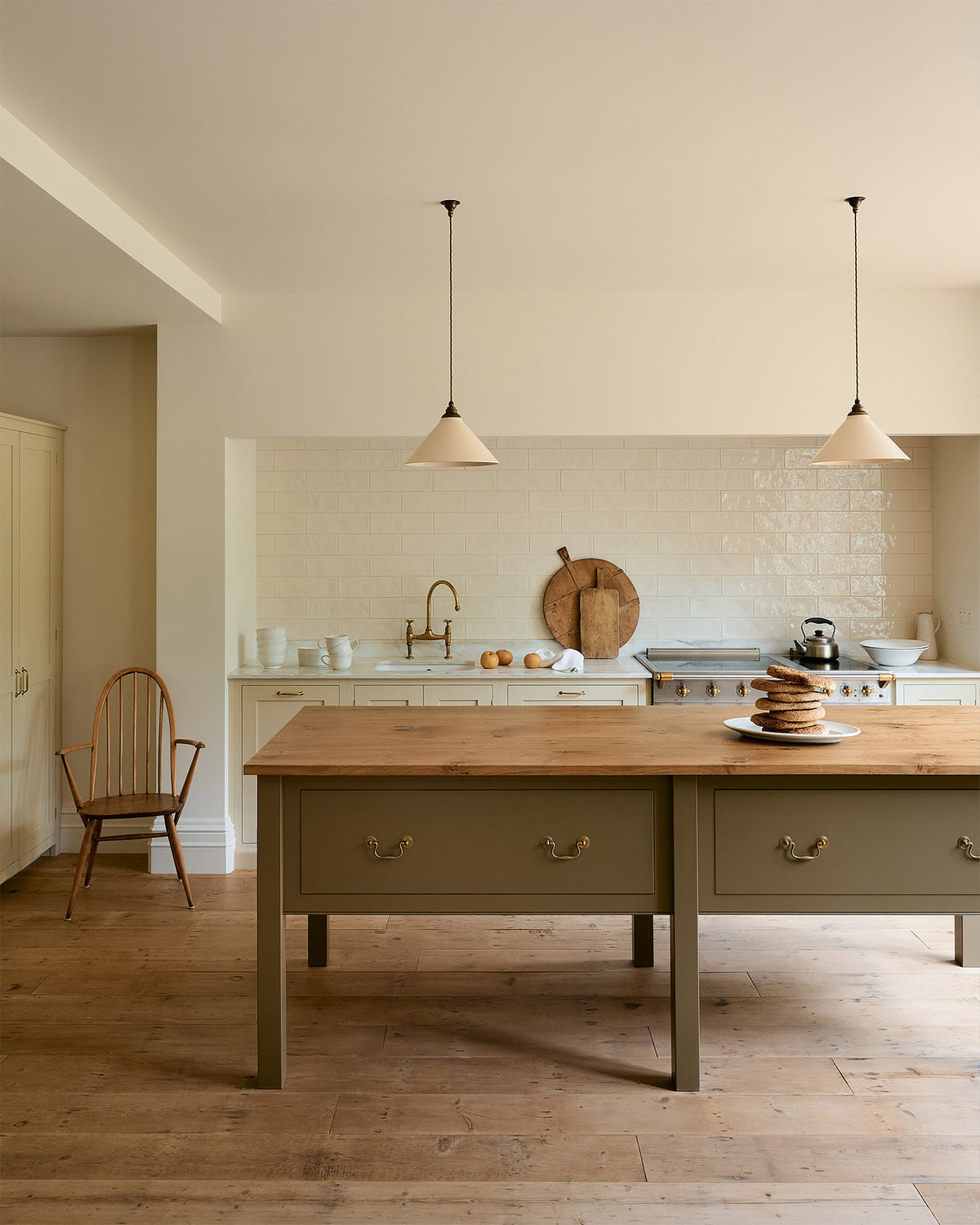 A light, clean, and simple shaker kitchen features a sink and cooker run nestled in a chimney breast, cream porcelain splash back tiles, wooden floorboards, and white walls. Cream cabinets complement an olive green Georgian-style prep table with deep drawers and a rustic oak worktop at the room’s centre, illuminated by two elegant creamware pendant lights.