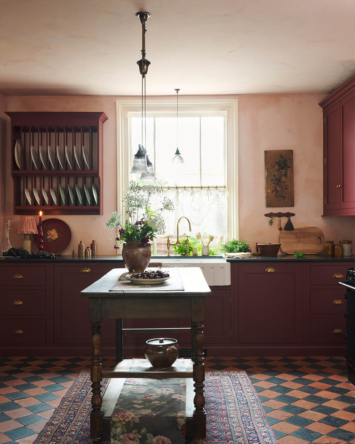 A warm and simply gorgeous refectory-red Shaker kitchen features a sink run with a black granite worktop, a wall cupboard, a small Heirloom plate rack and a wall cupboard. Beautifully decorated with vintage furniture, artisan ceramics, original botanical illustrations, reeded glass pendant lights, and a wild floral arrangement, it creates a humble and endearing space.