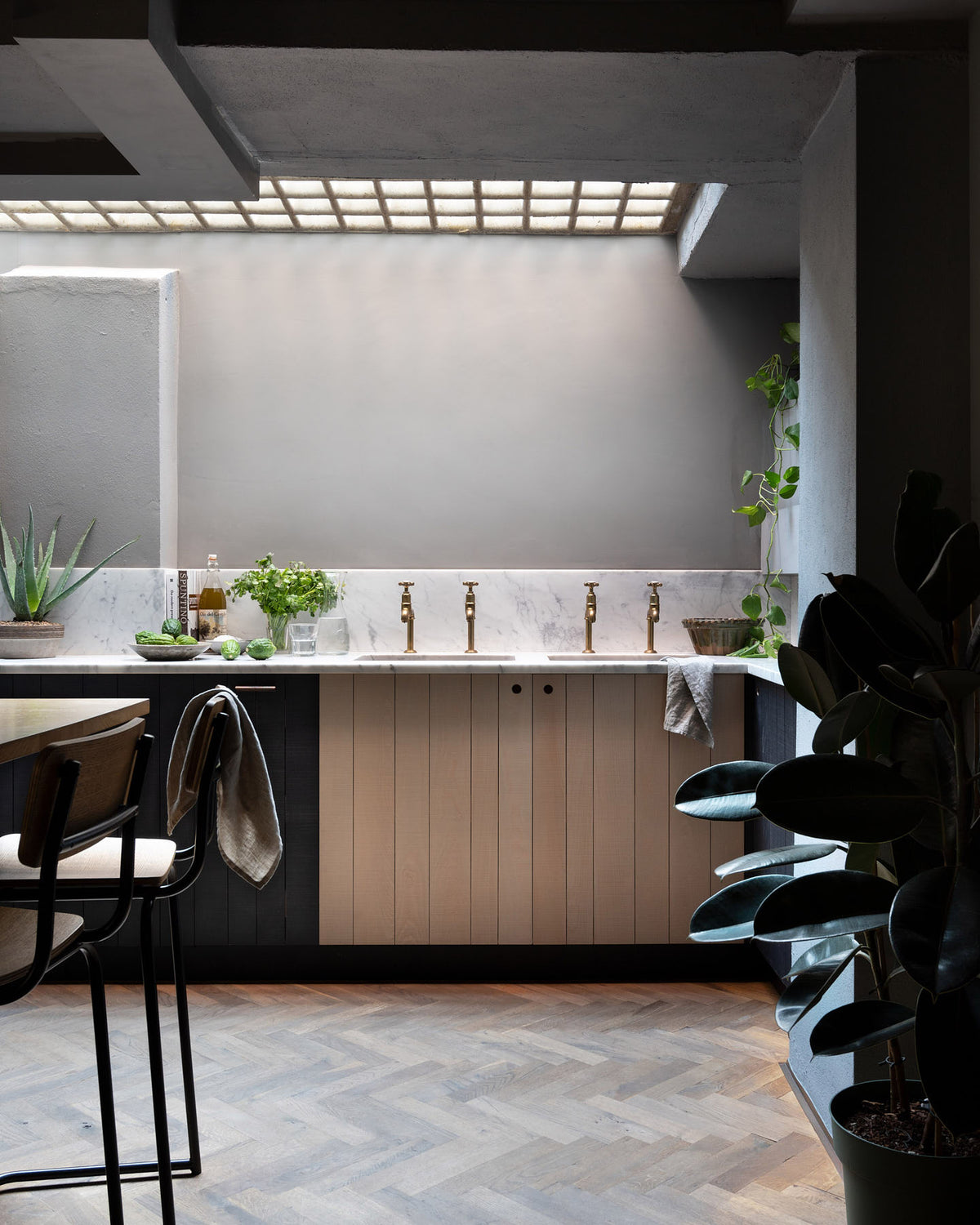 A chic and simple Sebastian Cox basement kitchen features a long sink run with two sets of copper sinks, aged brass Mayan taps, a Carrara marble worktop, glazed wall cupboards, a large kitchen island with a solid oak worktop, and a breakfast nook. This kitchen creates a cool and atmospheric space, beautifully decorated with luscious green house plants, artisan ceramics, a carafe of water, a drinking glass, a plumber’s stool, and a simple pendant light.
