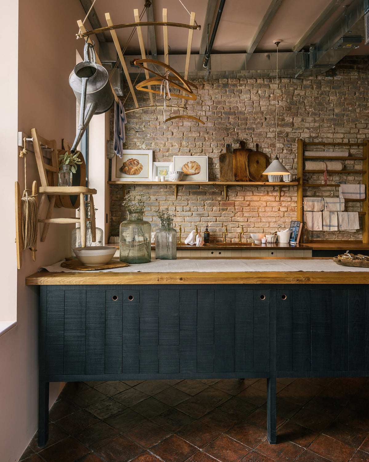 A calm and simple Sebastian Cox kitchen features a long sink run with two sets of copper sinks and aged brass Mayan taps, a copper worktop, a shelf, and a preptable with a solid oak worktop. Adorned with vintage items, artisan ceramics, wooden chopping boards, framed illustrations, tea towels, and wildflowers in large glass containers, this kitchen creates a chic New York industrial space that celebrates a love of natural materials.