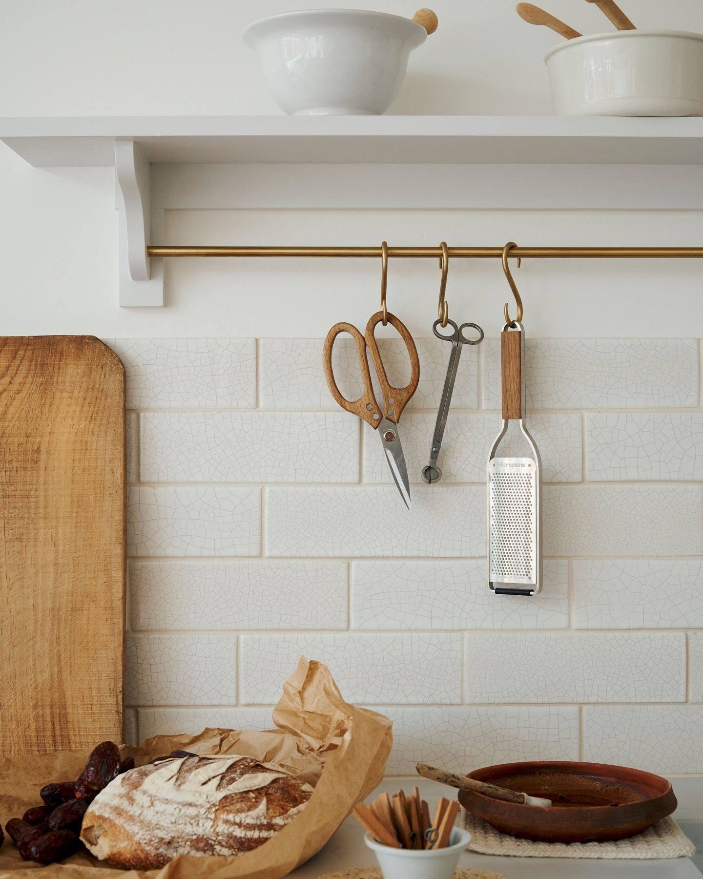 A close-up view of an assortment of kitchen utensils hanging from a brass rail in front of white metro wall tiles with delicate crazing.