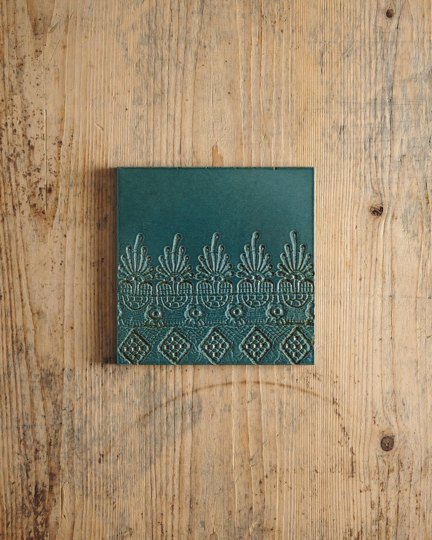 A glossy teal square tile with a decorative lace pattern on a rustic wooden surface.