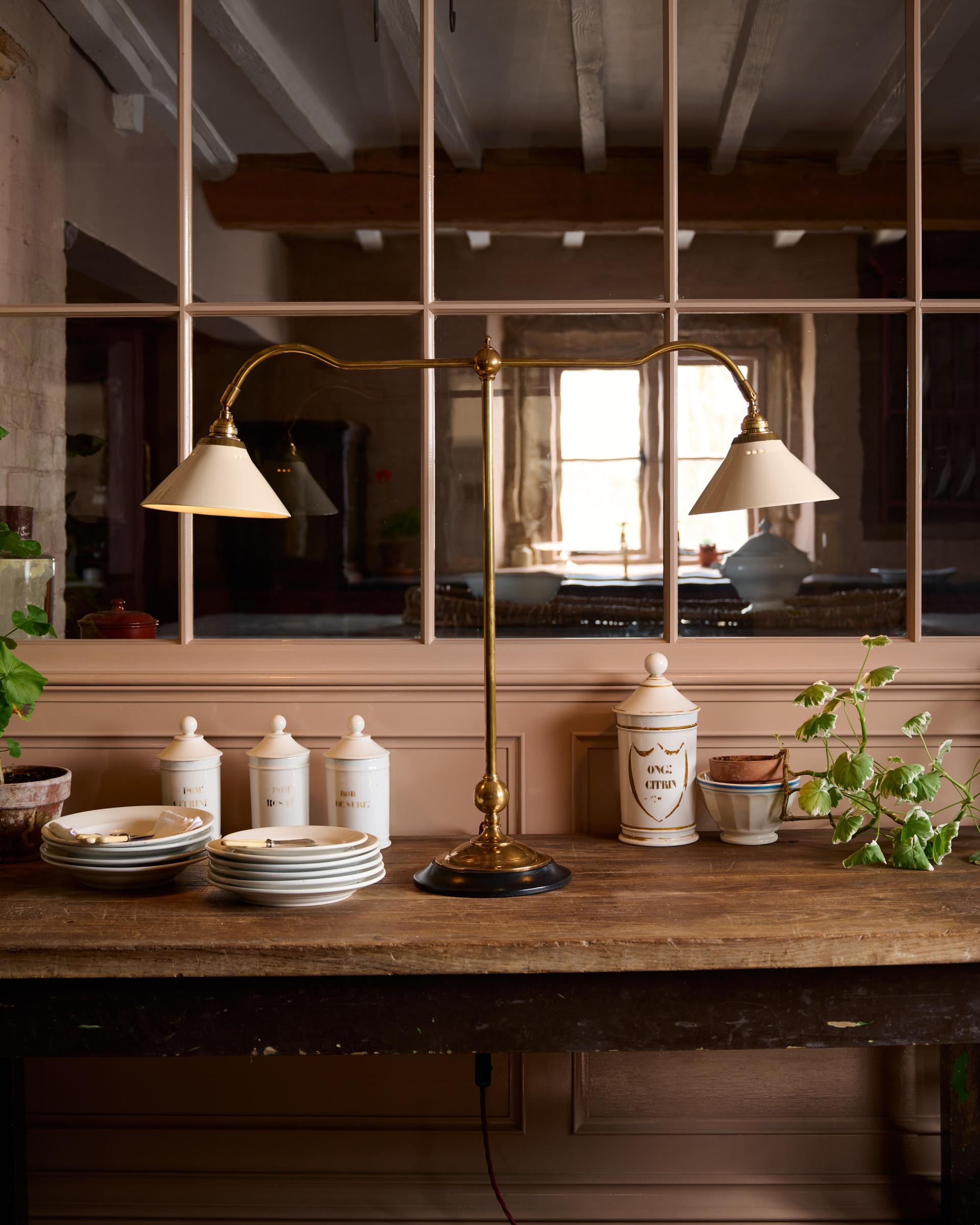 A straight-on view of a Victorian-inspired desk lamp with a solid brass foot, stand, two curved arms and two small cream glazed ceramic light shades on a table in front of a glazed partition.