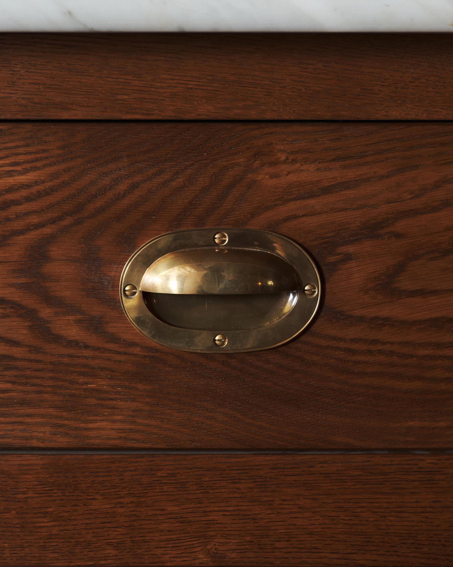A straight-on view of a Victorian-inspired oval-shaped cup handle with an authentic and warm burnished brass finish and a recessed backplate, fitted to a dark stained oak drawer front.