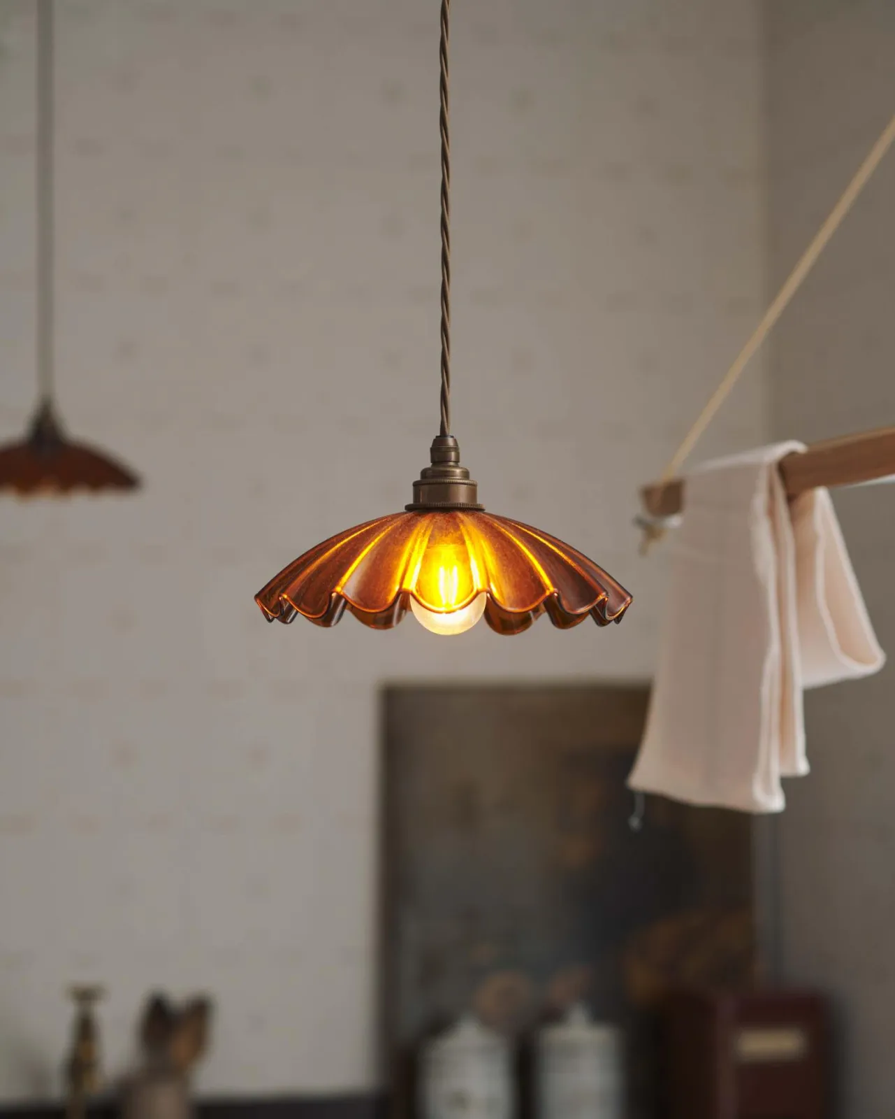 A close-up view of a small frilly amber glass pendant light with brass fittings and brown twisted fabric flex cord. The light is on and shining through the tranlucent glass.