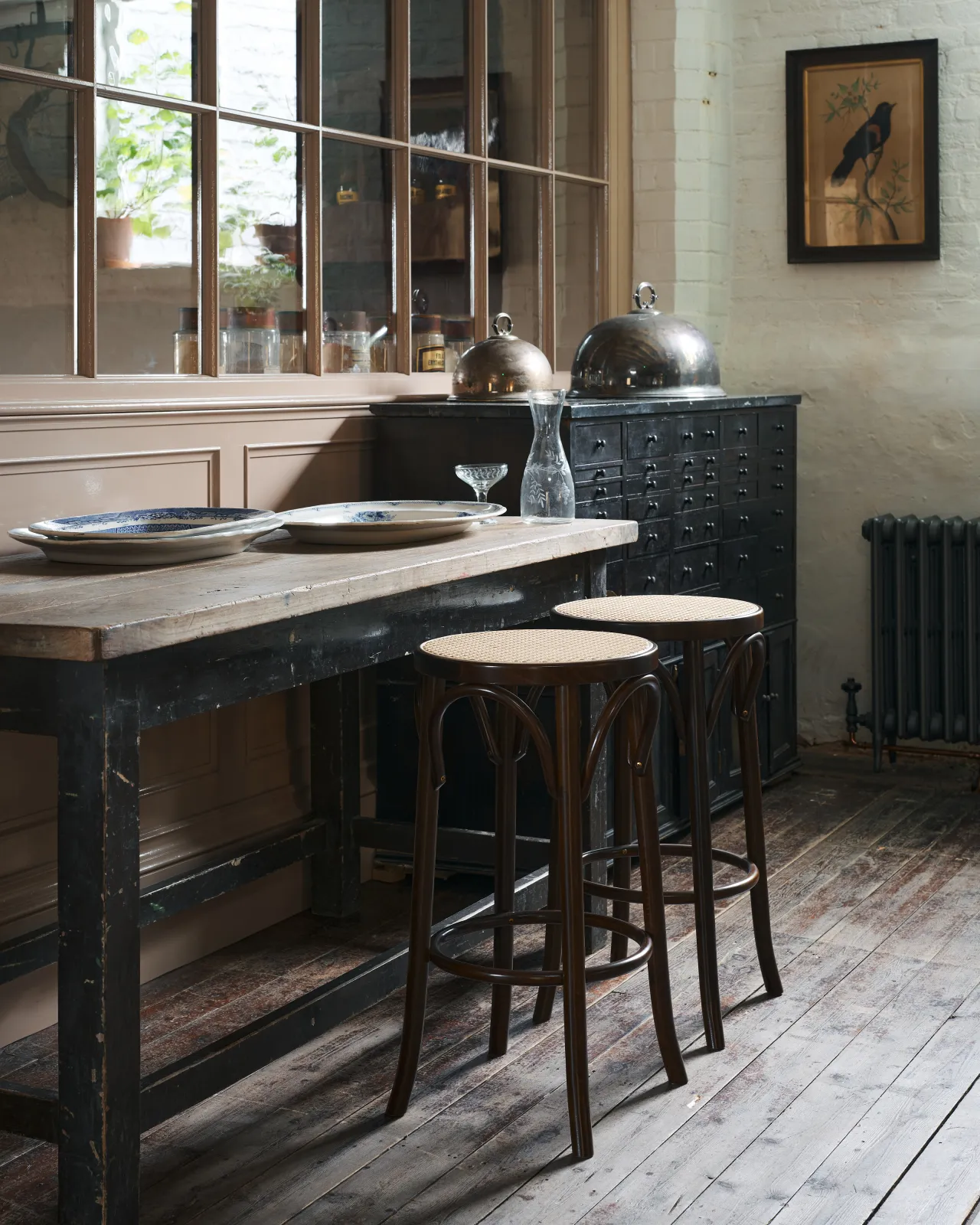 An oblique view of two café-style bar stools with dark stained frames and light woven ratten seats by a vintage table.