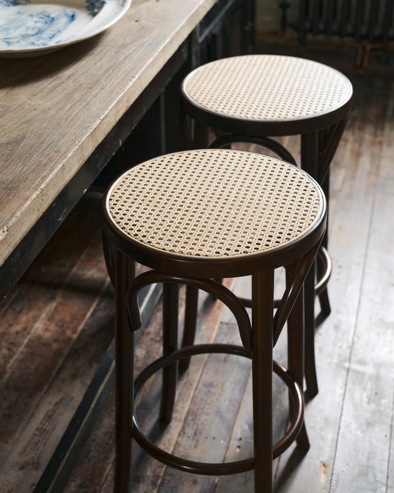 A close-up oblique view two café-style bar stools with dark stained frames and light woven ratten seats.