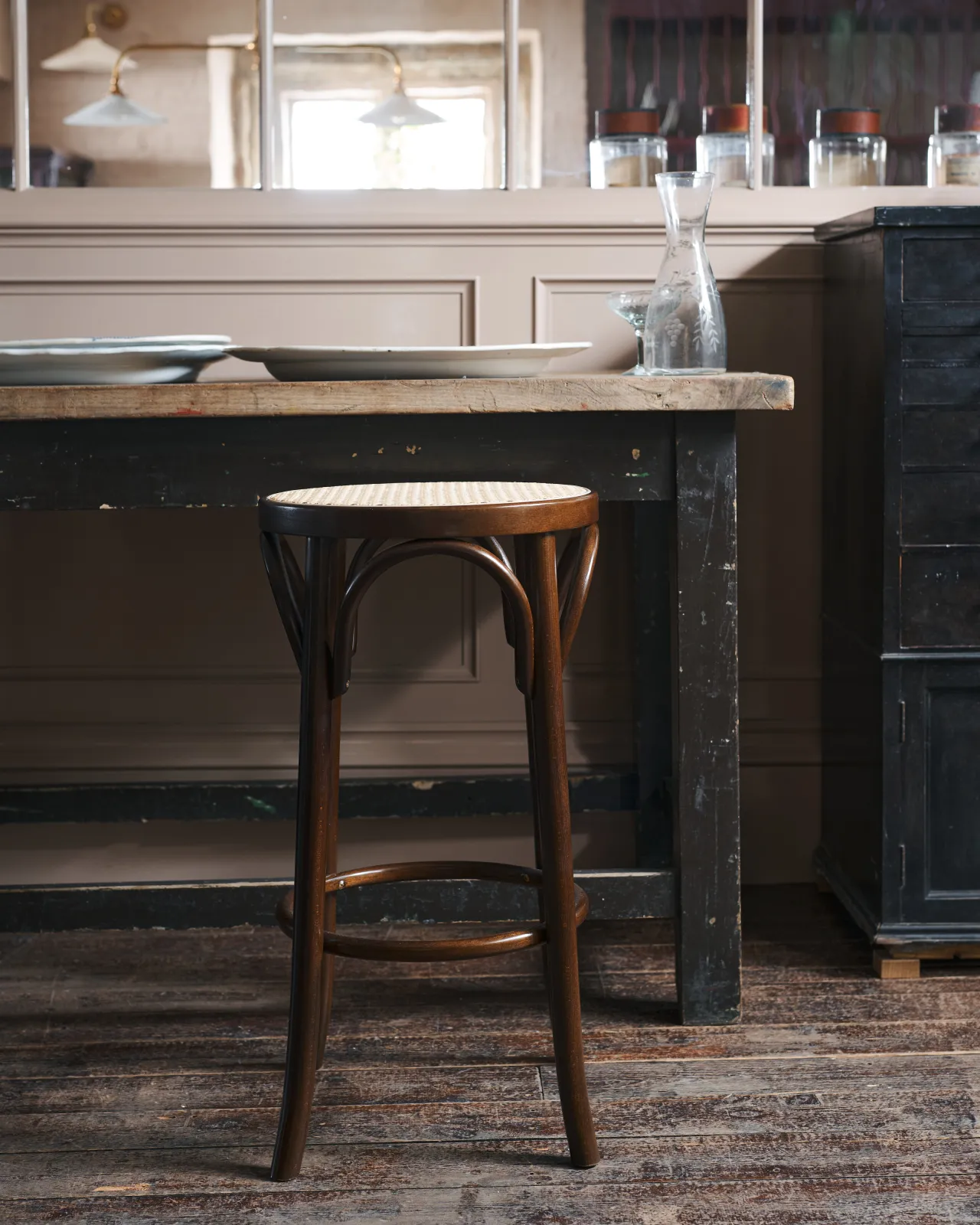 A straight-on view of a café-style bar stool with a dark stained frame and light woven ratten seat by a vintage table.