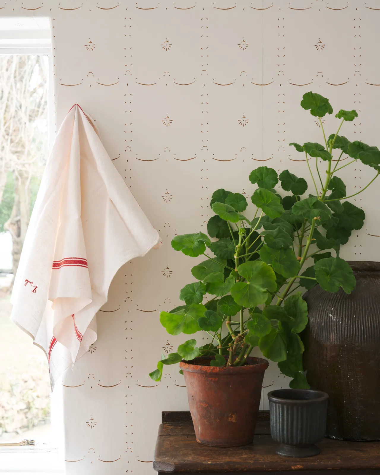 A delicate hand-painted wallpaper around a window with lush geraniums and a vintage tea towel hanging nearby.