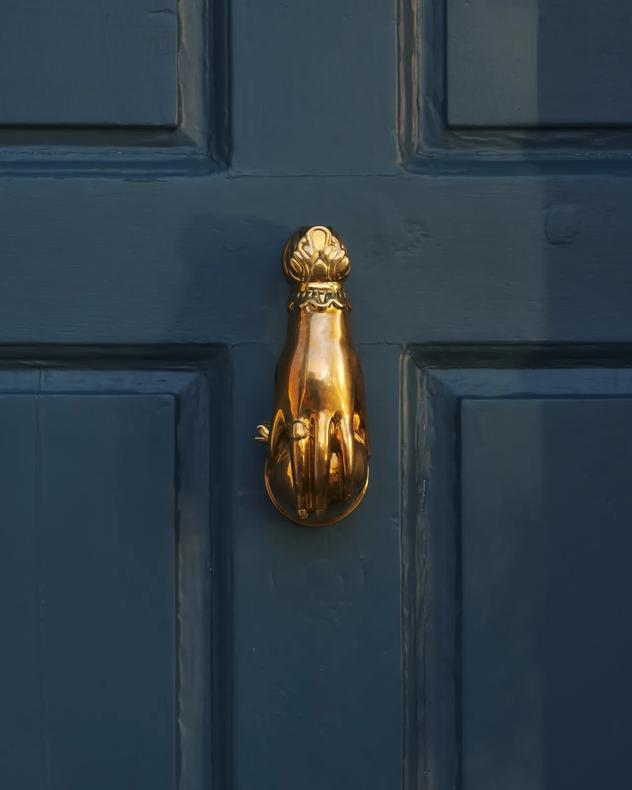 A close-up straight-on view of a decorative brass door knocker, shaped like a hand holding a pomegranate and fitted to a blue painted door.