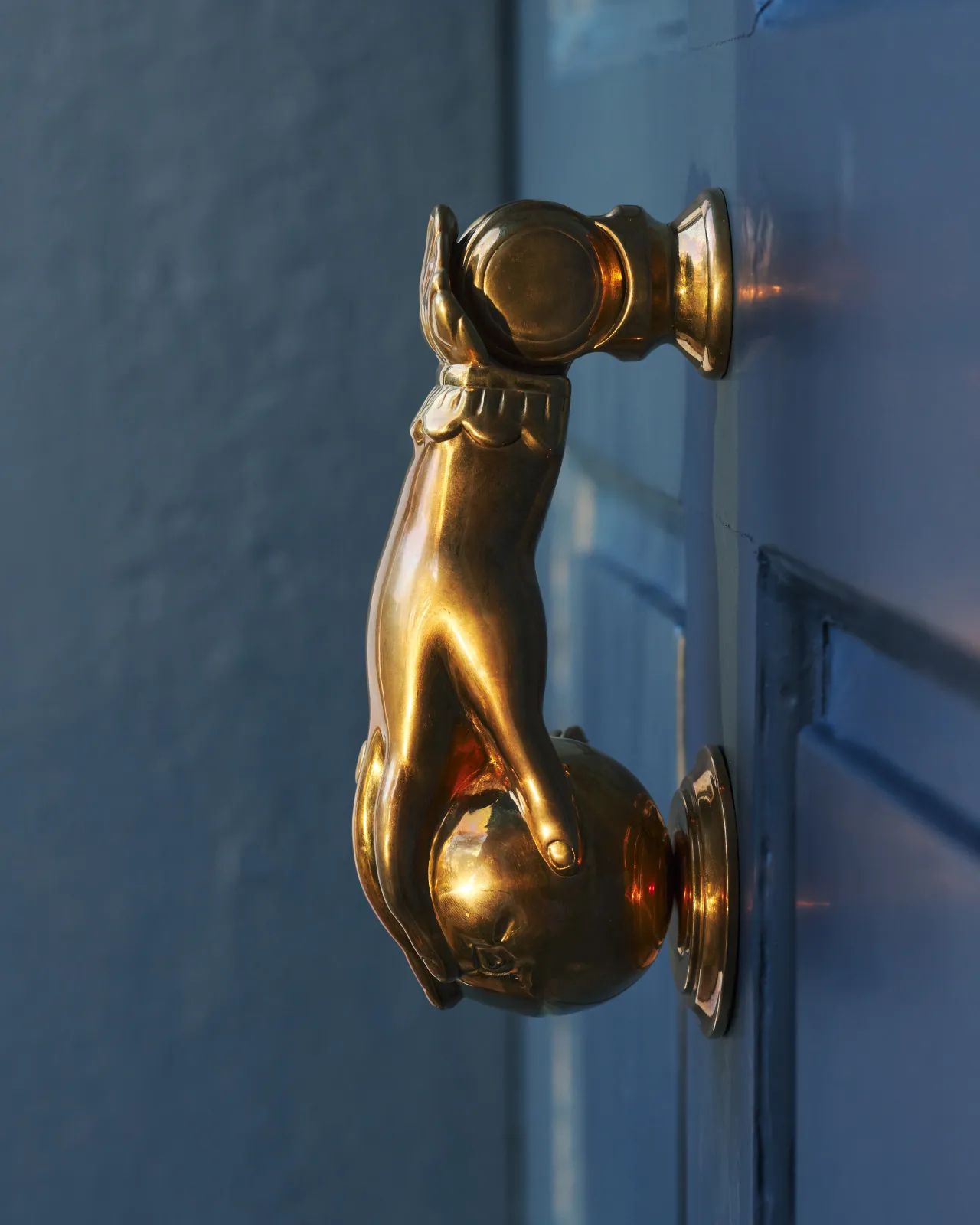 A profile view of a decorative brass door knocker, shaped like a hand holding a pomegranate and fitted to a blue painted door.