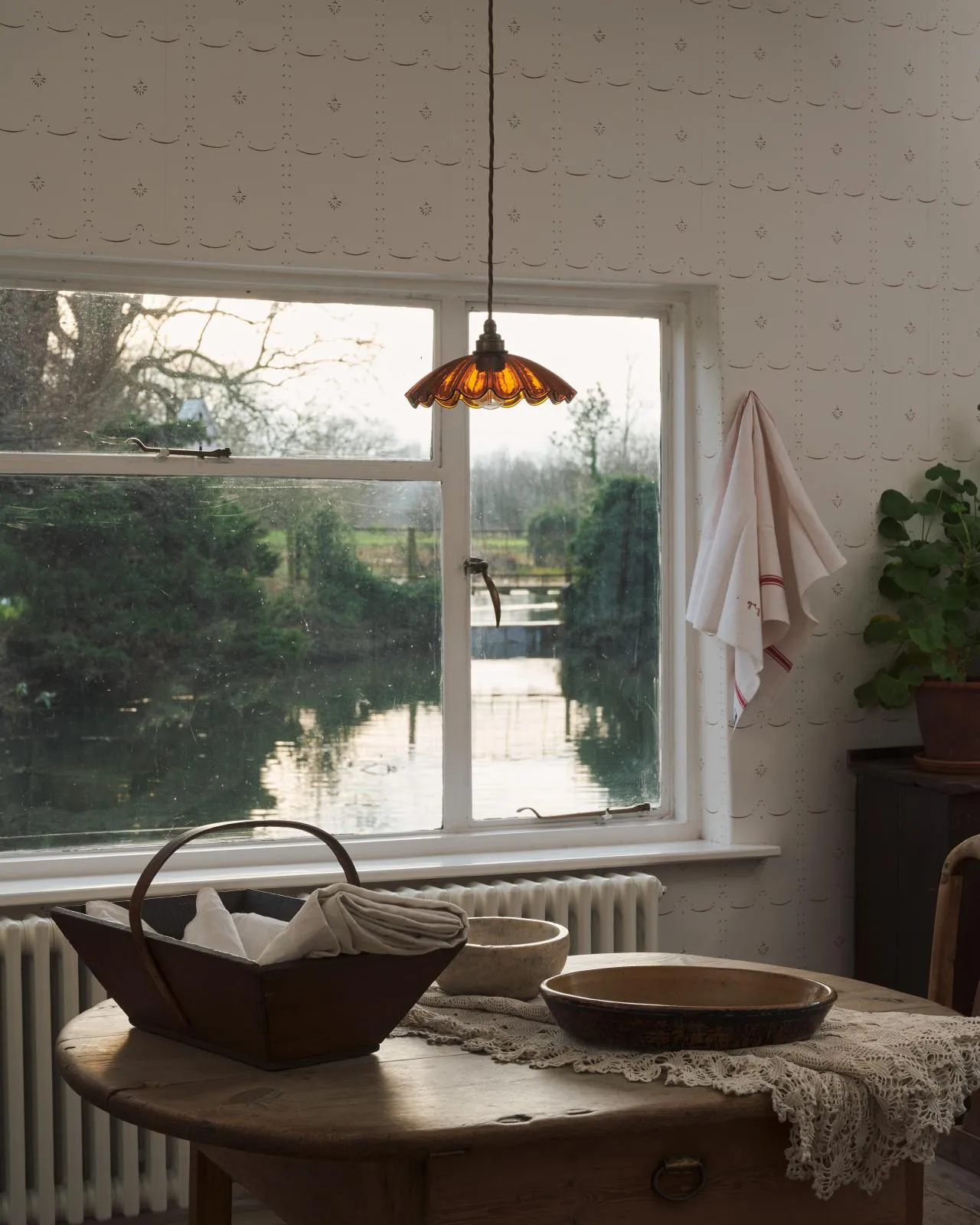 A small frilly amber glass light shade with brass fittings and brown twisted fabric flex cord hanging in front of a large window with views over a millpond and lush English countryside.