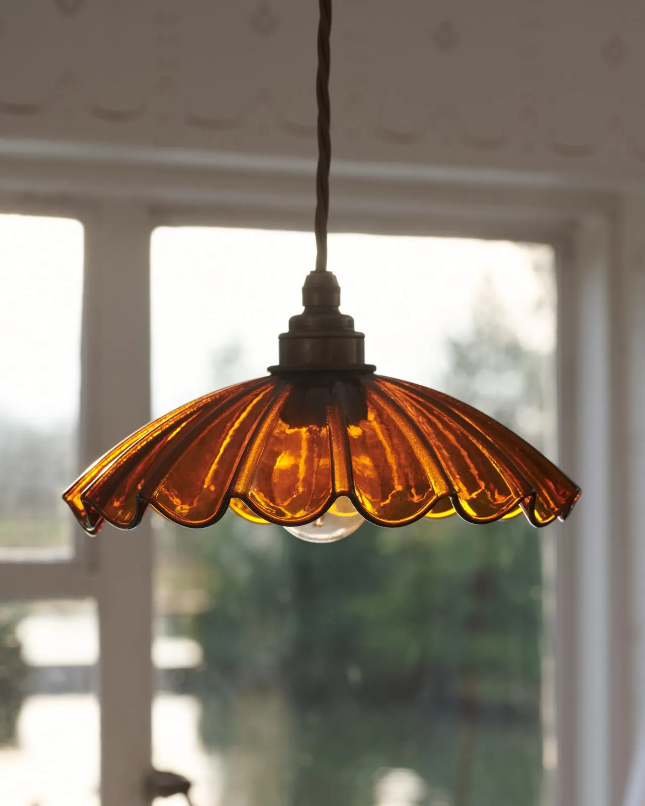 A close-up view of a small frilly amber glass light shade with brass fittings and brown twisted fabric flex cord. Light from a window behind is shining through the translucent glass.