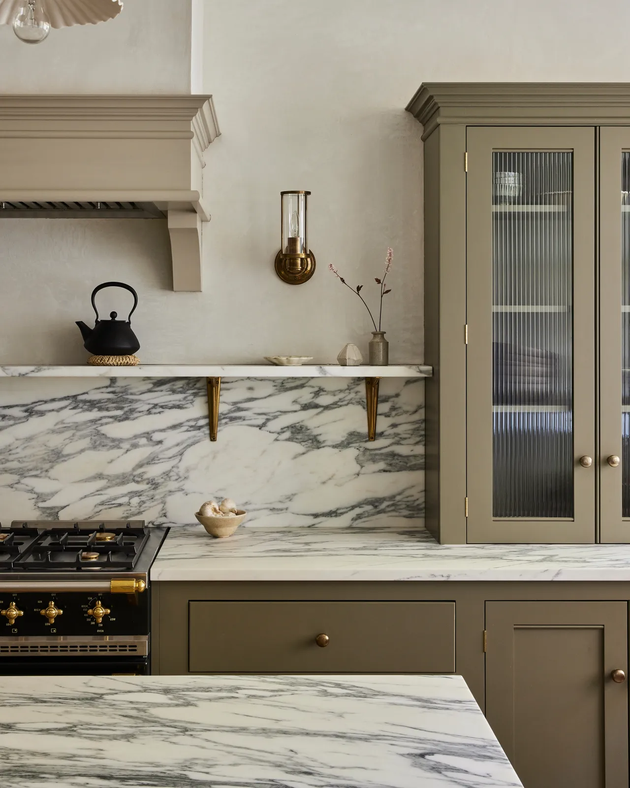 A classic contemporary kitchen with beige painted cupboards, reeded glass, Arabescato marble worktops, splashback and shelf, held up with classic brass utilitarian shelf brackets.