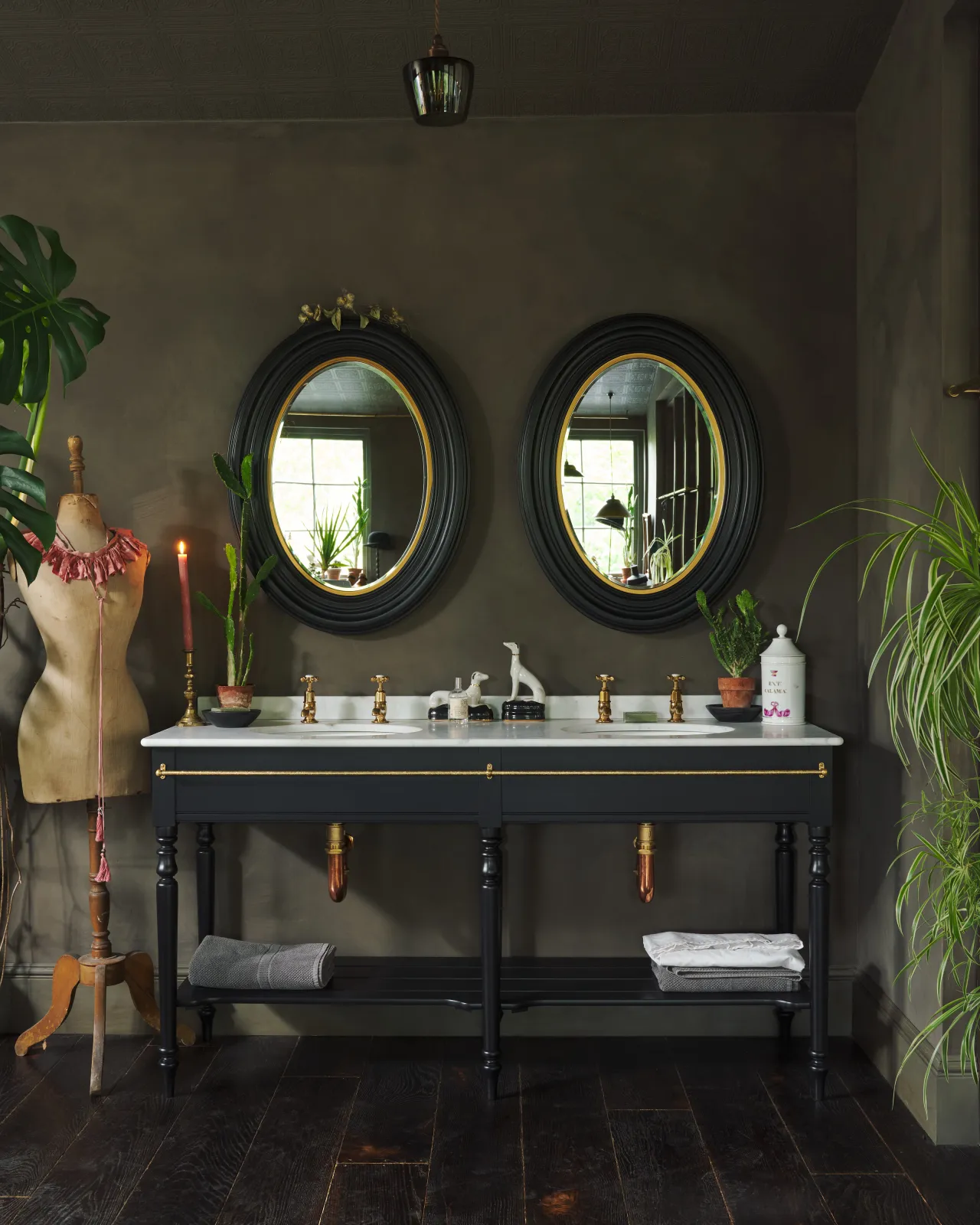 A straight-on view of a grand black Victorian-inspired washstand with two basins and two sets of brass pillar taps. Two black-framed oval mirrors sit above the basins, beside a large monstera plant and eclectic styling with vintage bric-a-brac.
