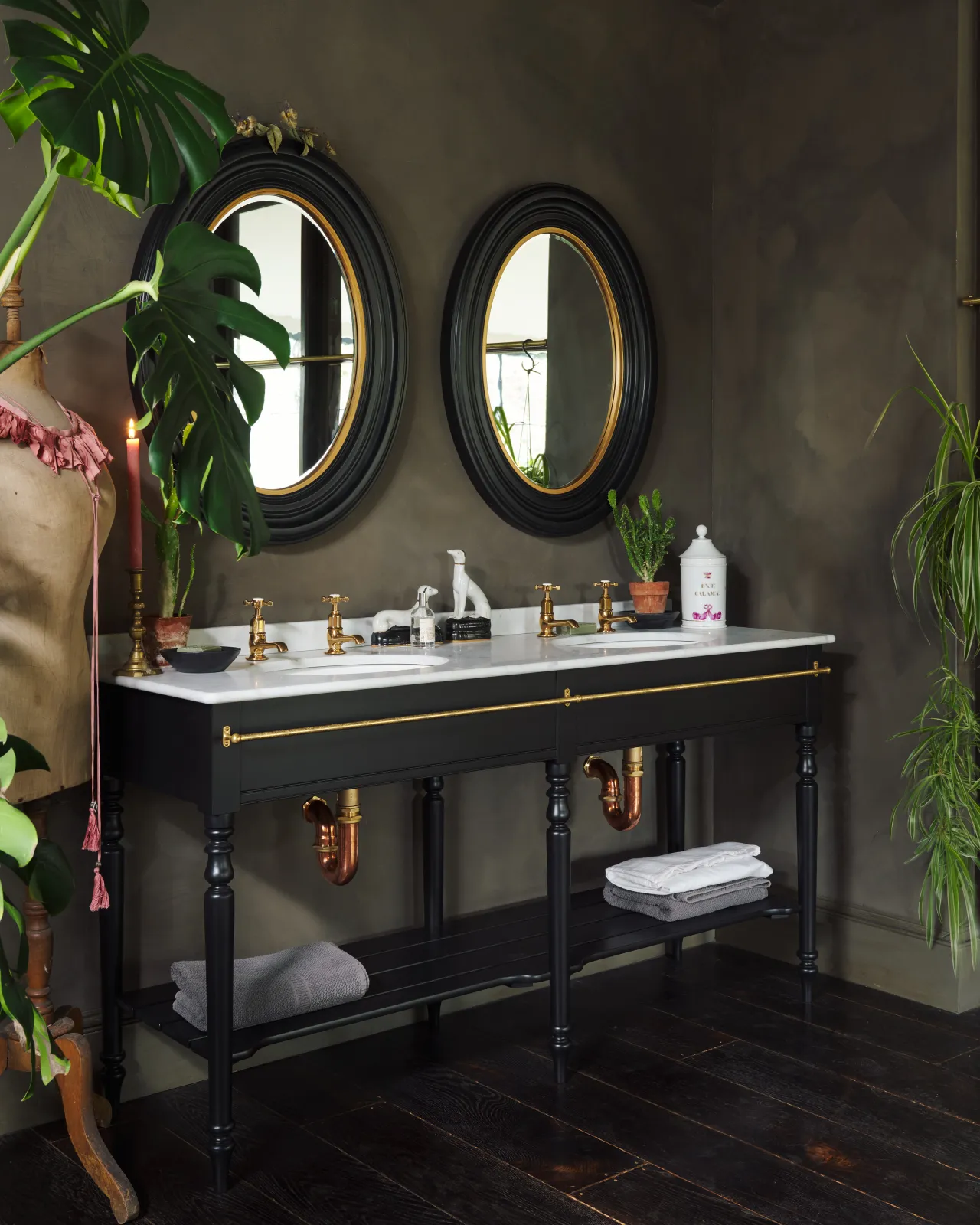 An oblique view of a grand black Victorian-inspired washstand with two basins and two sets of brass pillar taps. Two black-framed oval mirrors sit above the basins, beside a large monstera plant and eclectic styling with vintage bric-a-brac.