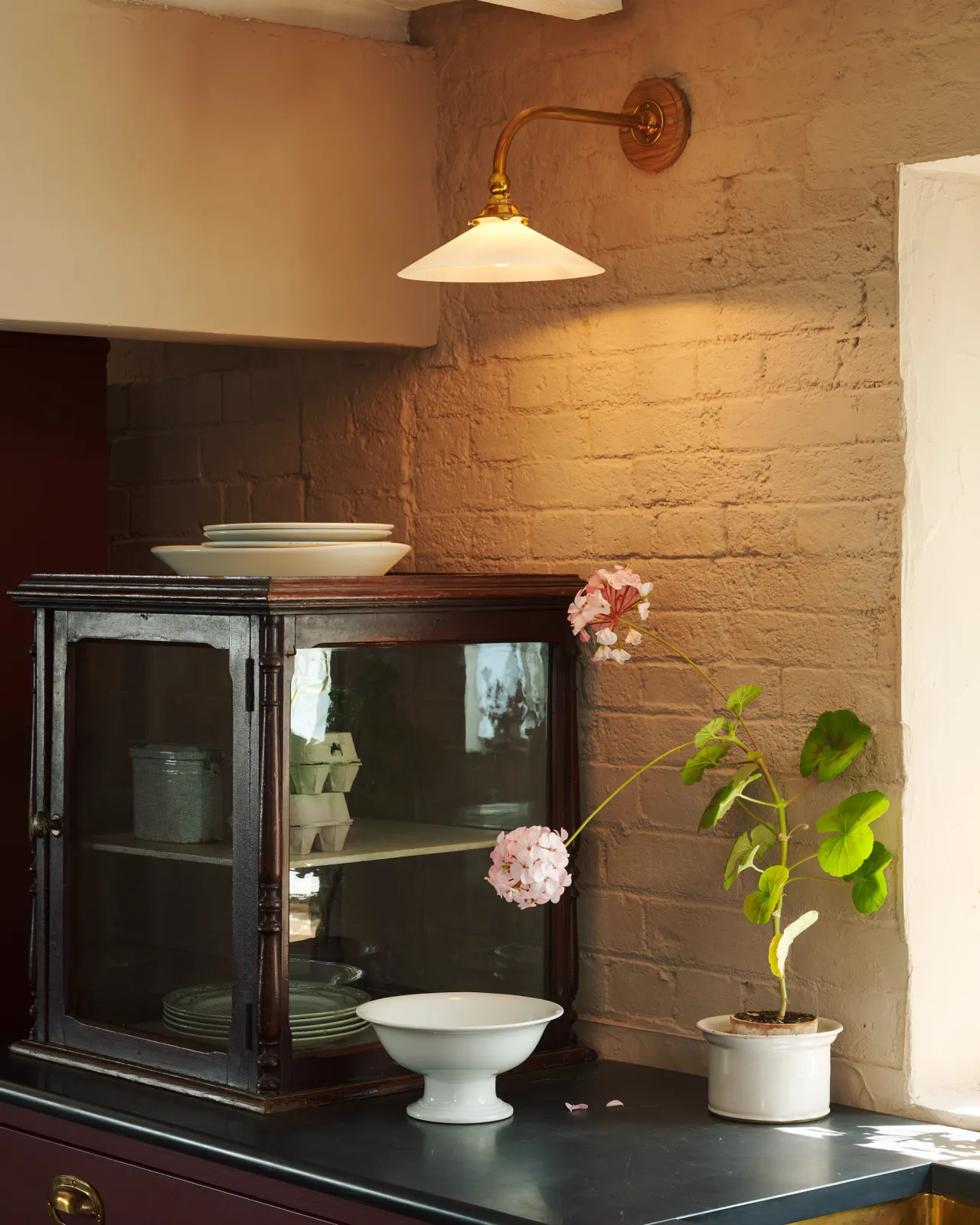 An oblique view of a small Victorian-inspired wall light with a brass arm and a milk glass shade fixed to a pink painted brick wall above a slate worktop and a vintage glazed cupboard.