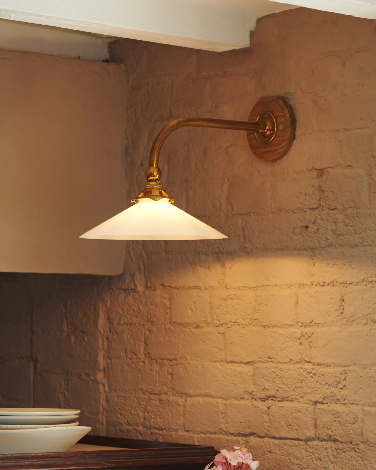 A close-up oblique view of a small Victorian-inspired wall light with a brass arm and a milk glass shade fixed to a pink brick wall.