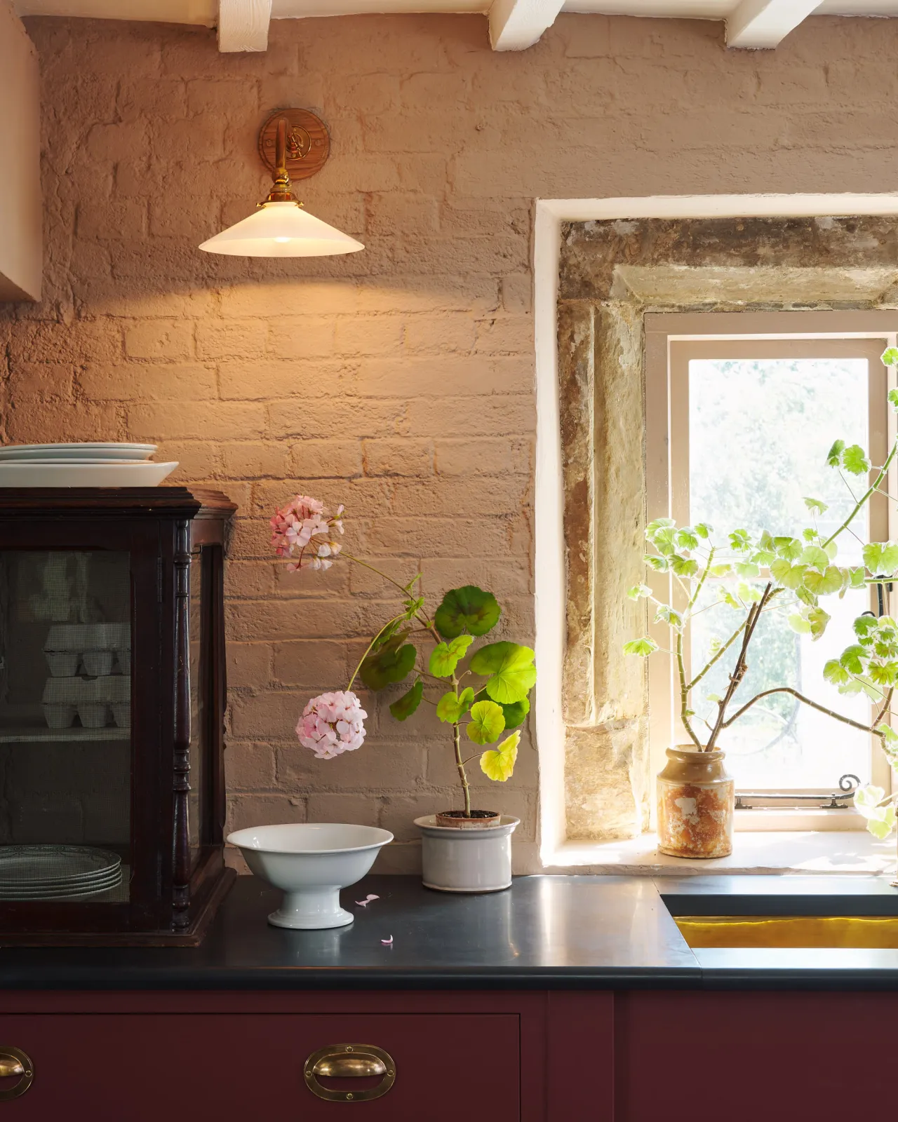 A straight-on view of a small Victorian-inspired wall light with a brass arm and a milk glass shade fixed to a pink painted brick wall above a slate worktop and red painted cabinetry.