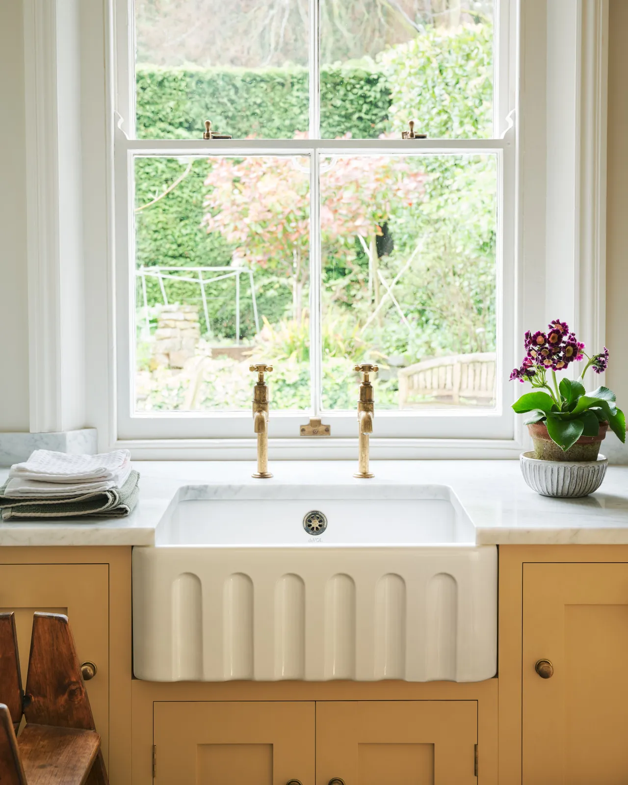 A close-up straight-on view of a small fluted ceramic Belfast sink in a yellow painted Shaker-style kitchen with aged brass bibcock taps.