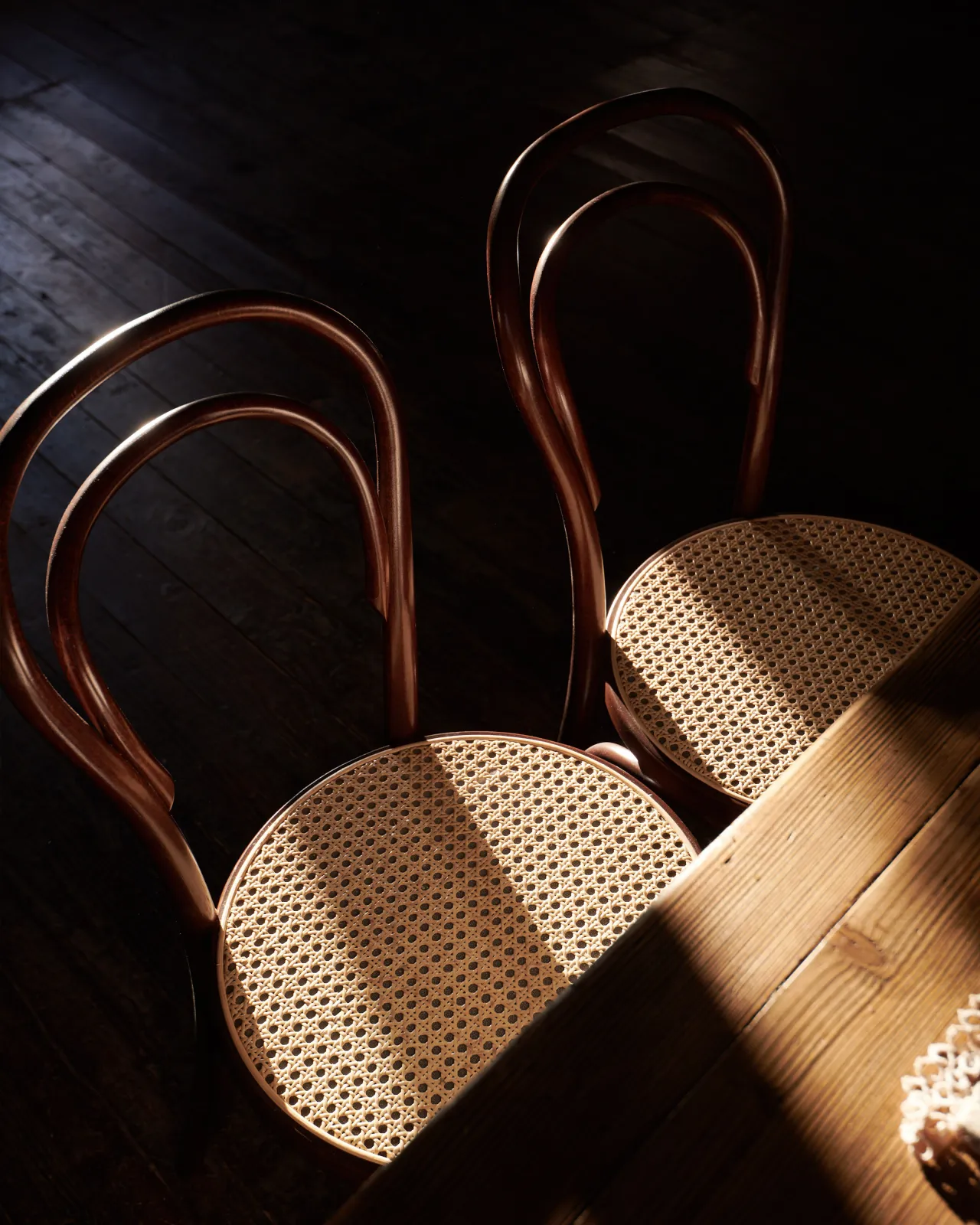 A dark room with two café-style bentwood chairs with dark stained frames and light woven rattan seat mesh.