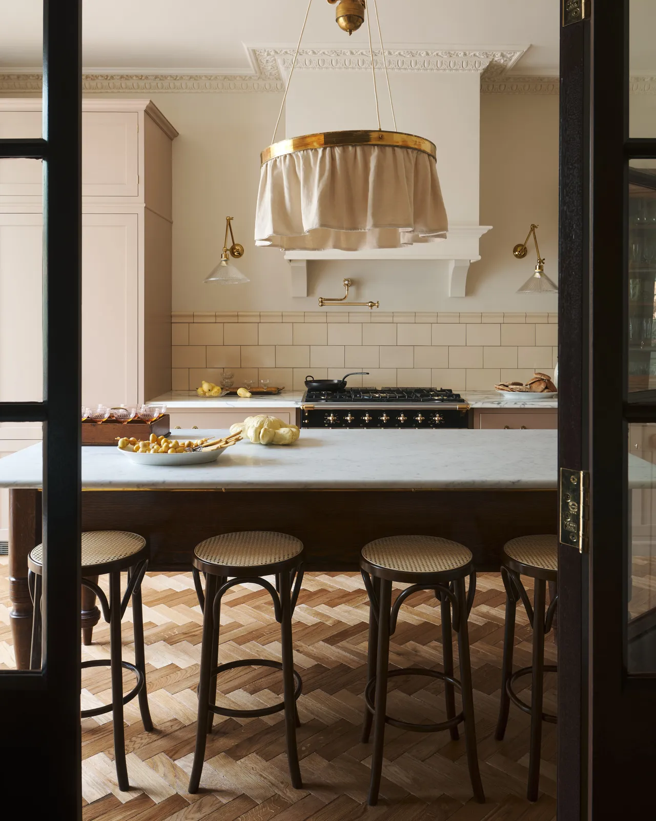 A straight-on view through a glazed partition into a smart bohemian kitchen with a row of café-style bar stools in front of a grand oak prep table with a Carrara marble worktop.