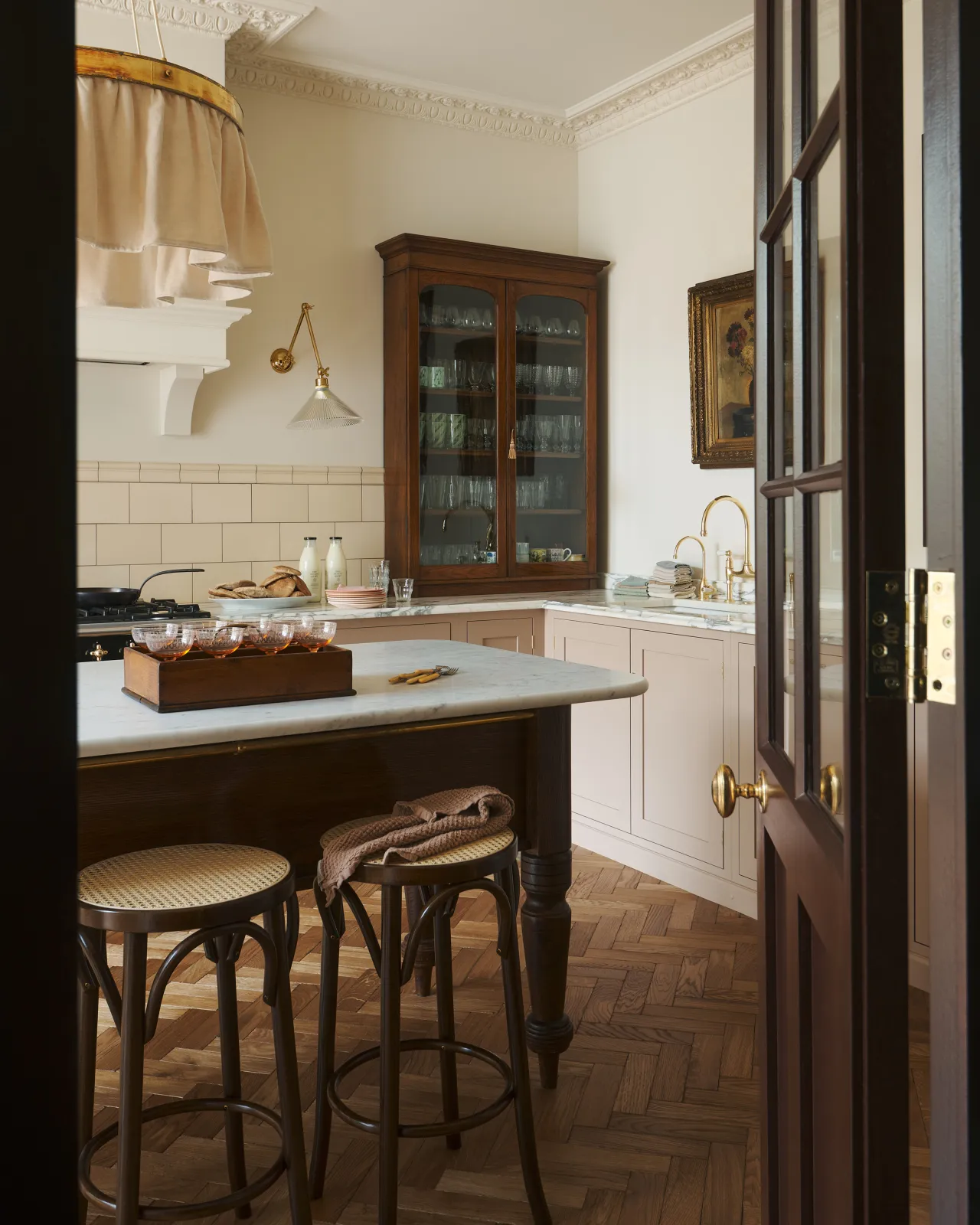 An oblique view through a glazed partition into a smart bohemian kitchen with a row of café-style bar stools in front of a grand oak prep table with a Carrara marble worktop.