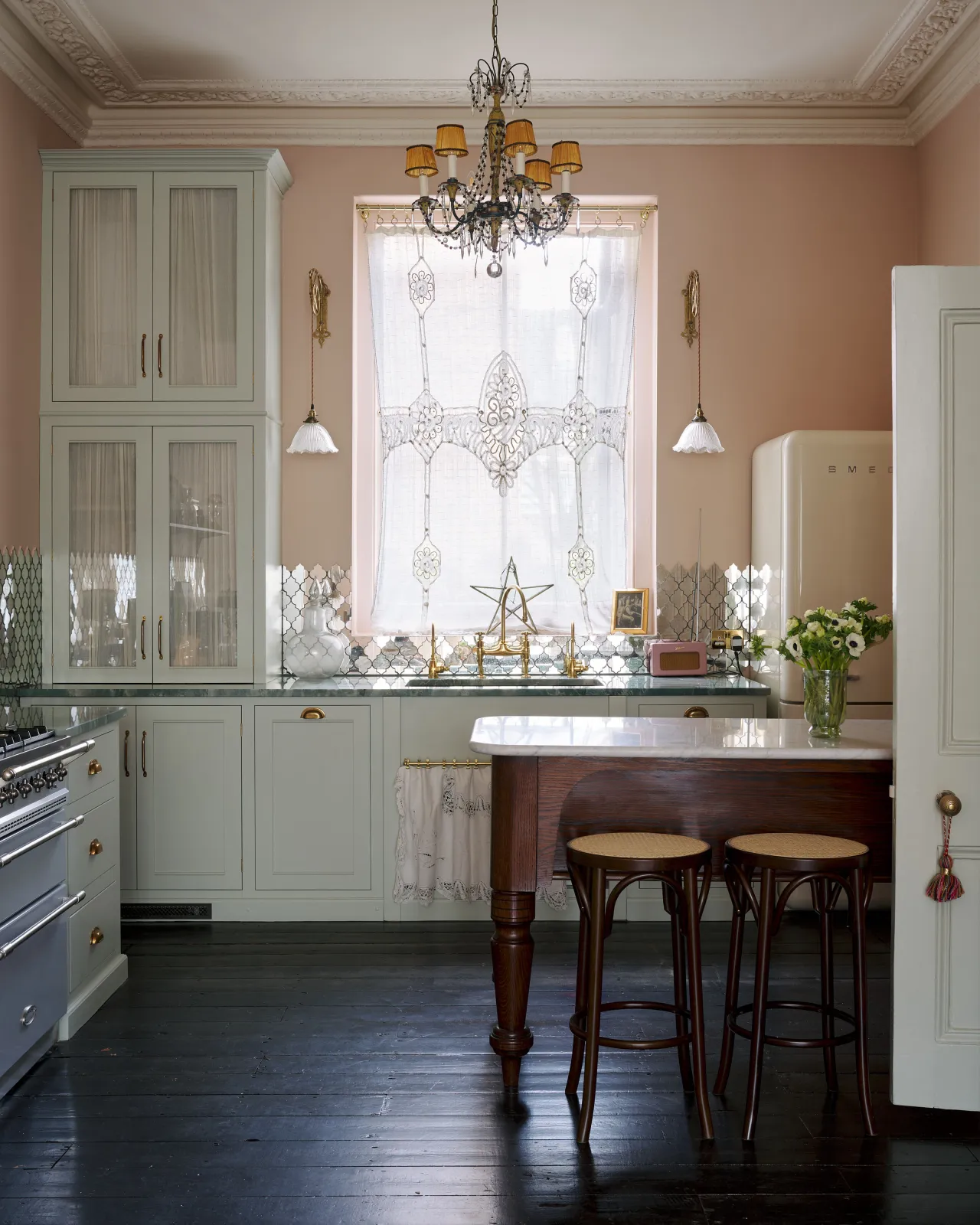 A bohmian-style kitchen with pink walls, duck-egg blue painted cabinetry, lace curtains and an oak prep table and two café-style bar stools with dark stained frames and light woven ratten seats.