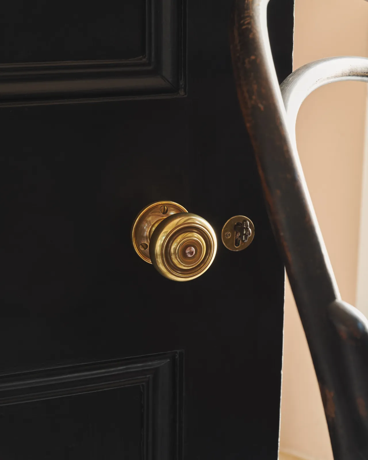 A close view of a traditional brass doorknob set against a deep black painted door, with a glimpse of a vintage bentwood chair beyond.