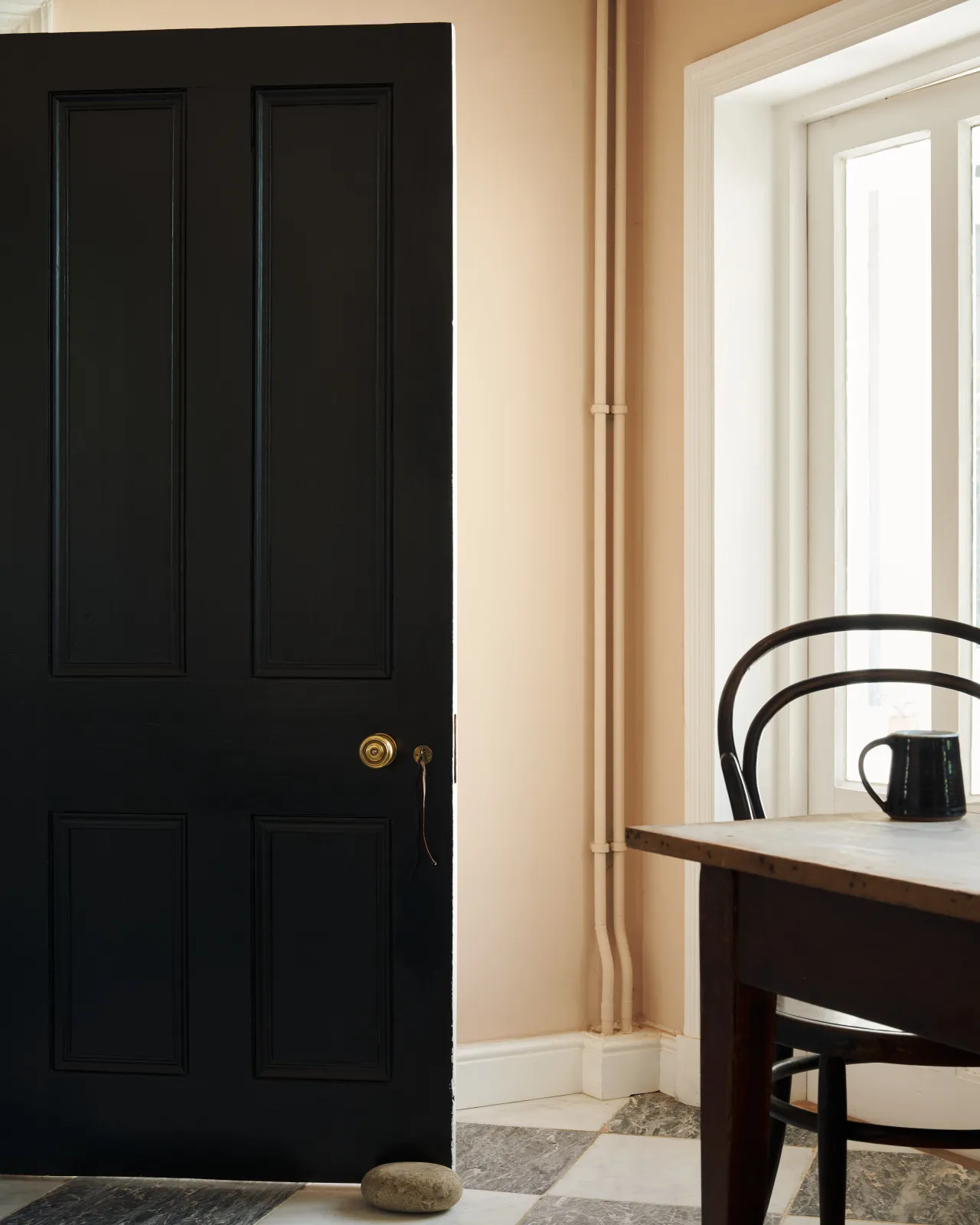 A full black-painted door in a soft blush-walled kitchen, fitted with a brass knob, quietly elegant beside a simple bentwood chair.