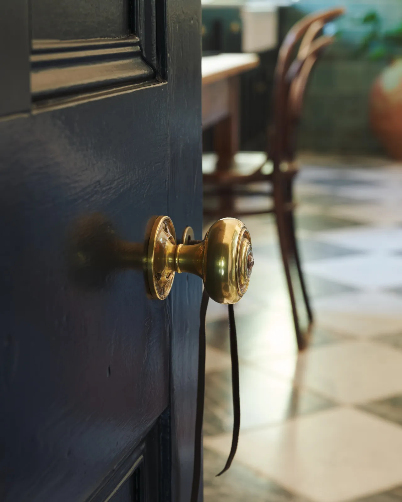 A partial open door revealing its brass knob, leading into a checkerboard-tiled kitchen with green tiled walls and bentwood chairs beyond.
