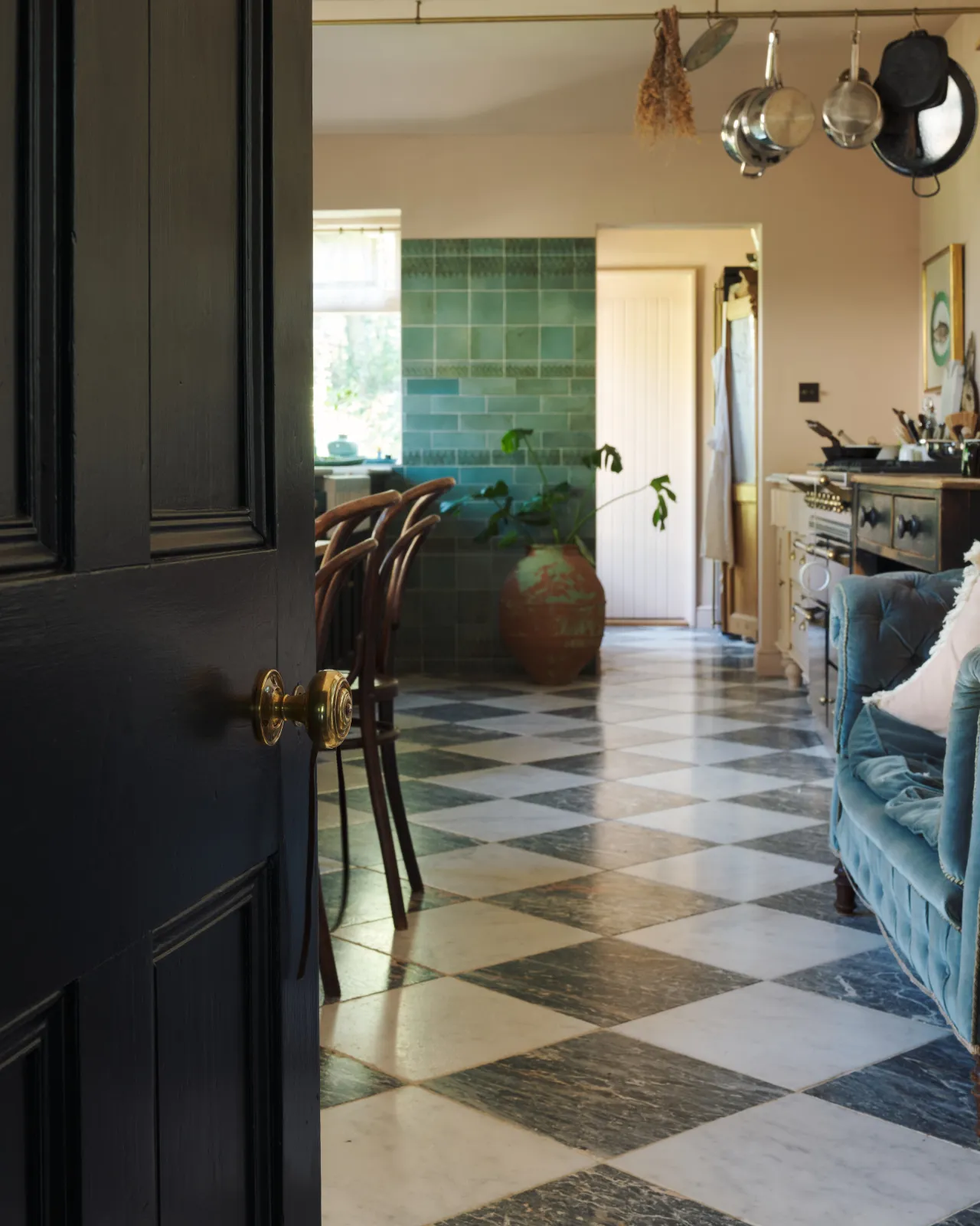 A partial open door revealing its brass knob, leading into a checkerboard-tiled kitchen with green tiled walls and bentwood chairs beyond.