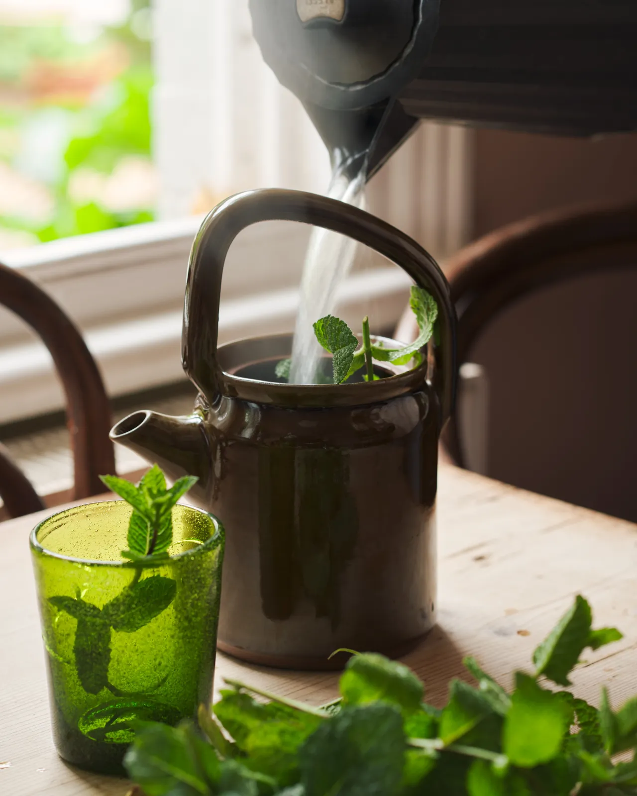 A black kettle pouring boiling water into a small mid-century-style ceramic teapot with a glossy amber glaze, surrounded by mint leaves and a small green glass.