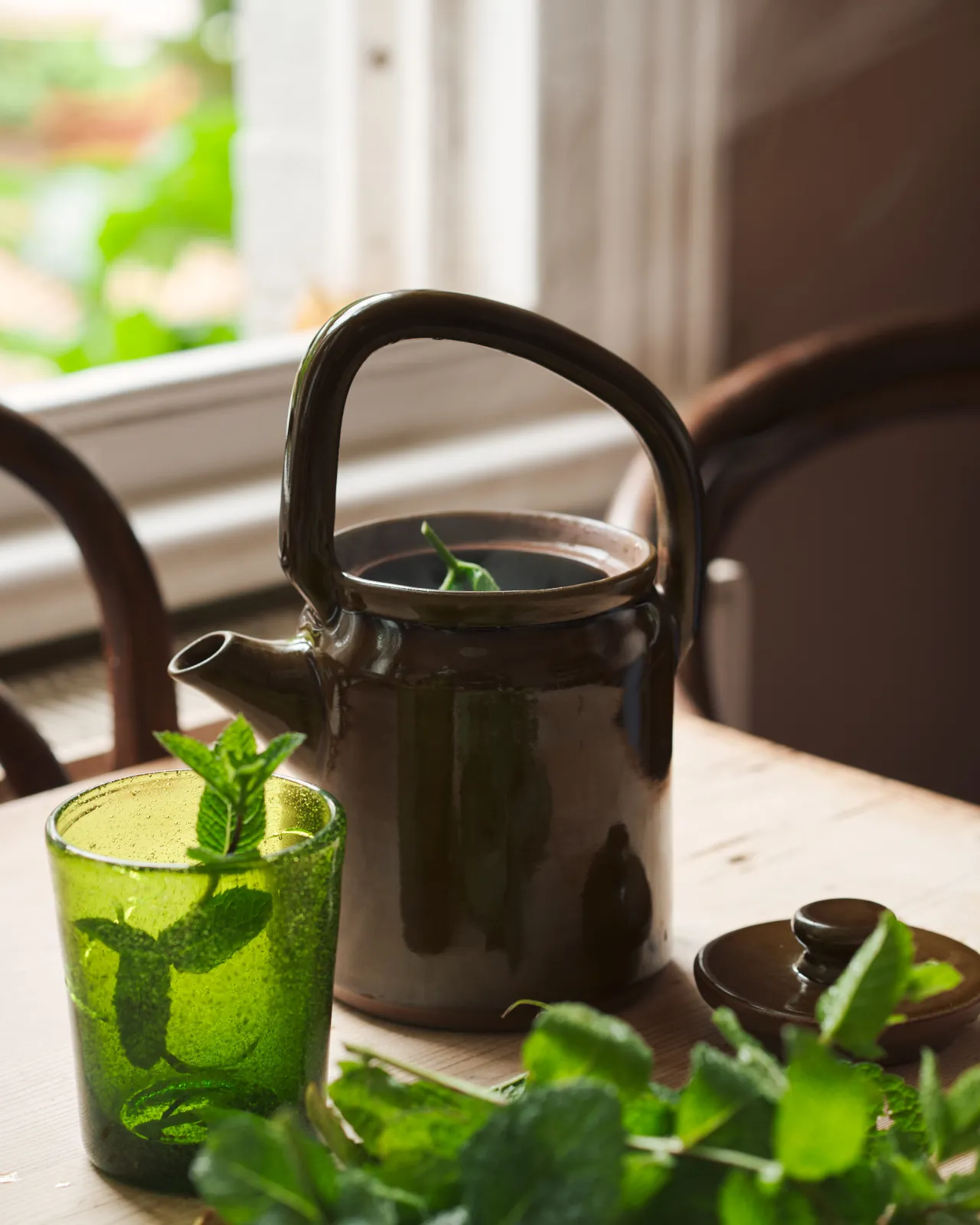 A small mid-century-style ceramic teapot with a glossy amber glaze, surrounded by mint leaves and a small green glass.